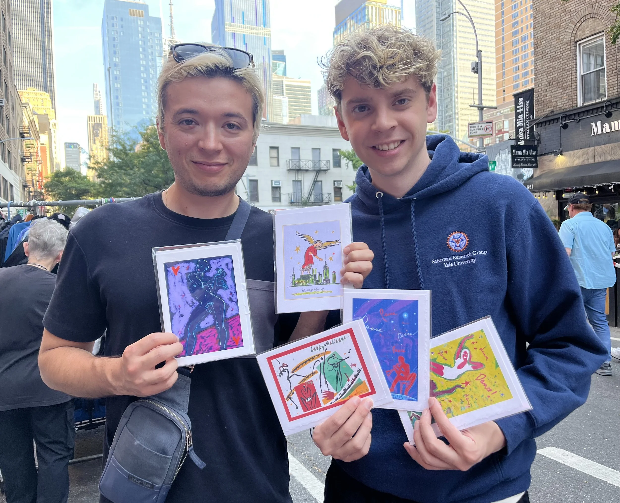 Two young men holding colorful JanetsArthouse cards in an outdoor city setting with tall buildings; one wears a black T-shirt, the other a navy hoodie with Yale University logo.