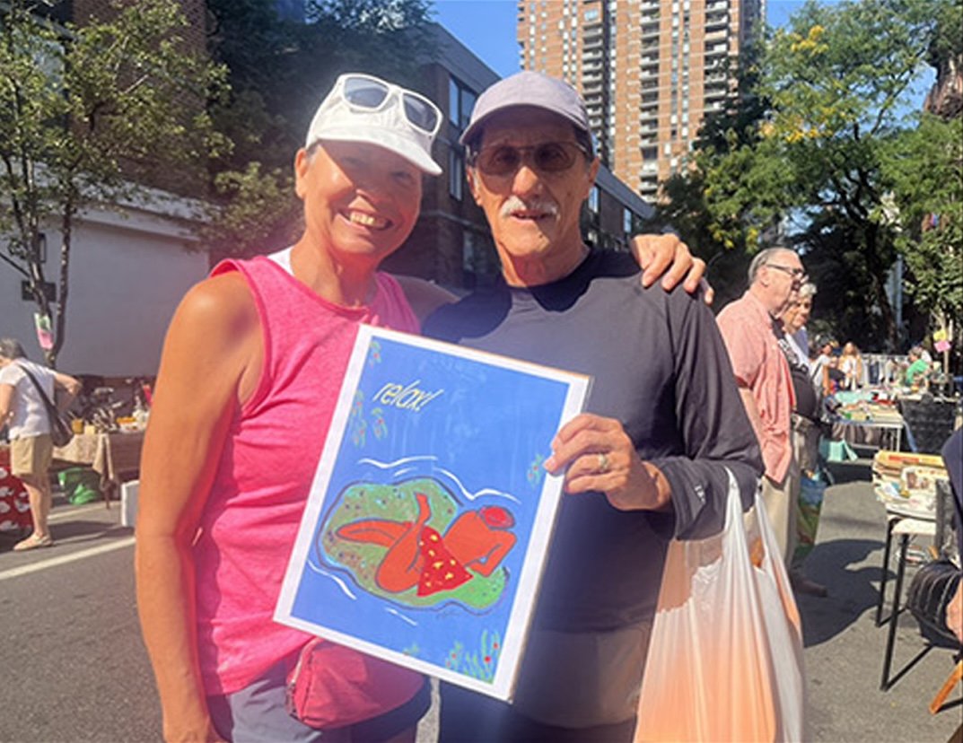 A smiling woman and an older man holding a colorful drawing of a red lobster with a red crab, on a city street during a market event, with tents and other people in the background.