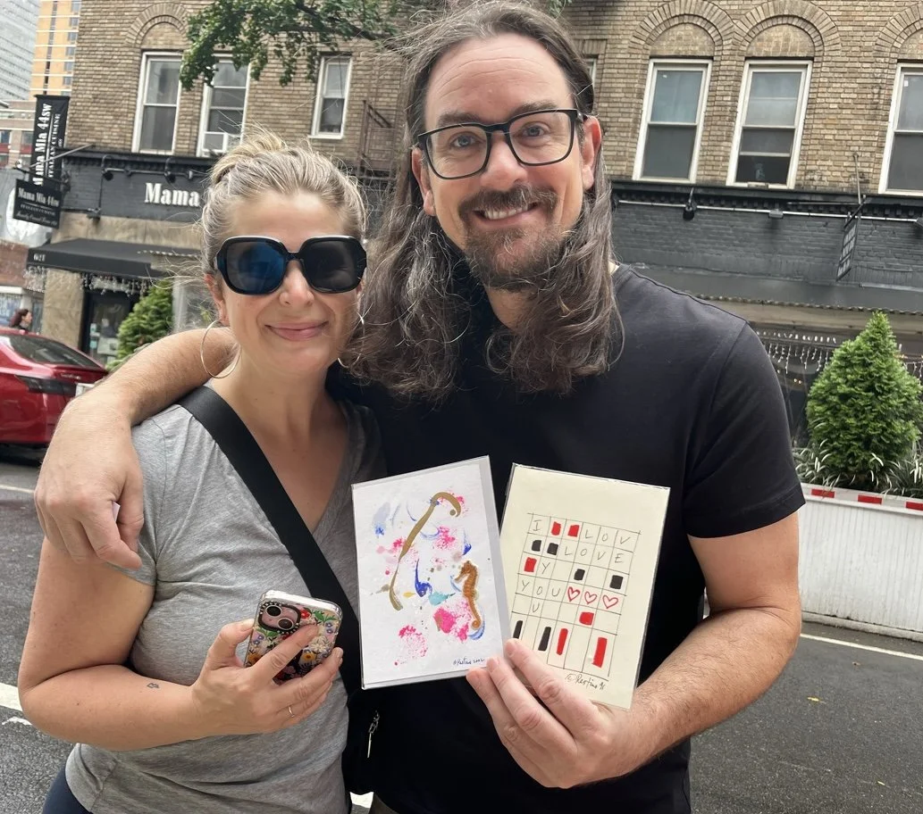 A man and woman smiling outdoors, holding artwork and a phone, with buildings and parked cars in the background.