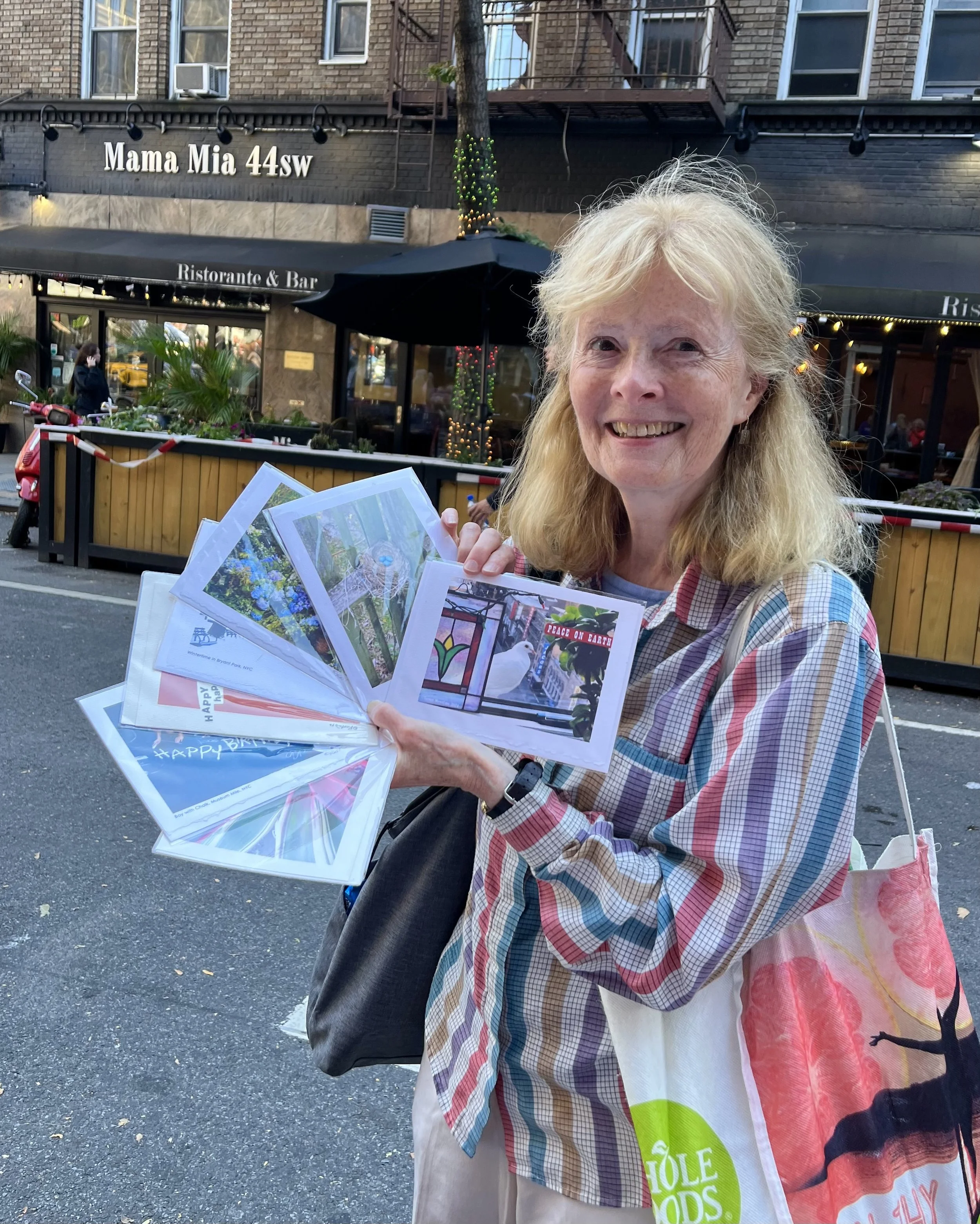 Smiling woman standing on city street holding a fan of greeting cards or prints, with a restaurant named 'Mama Mia 44 SW' in the background.