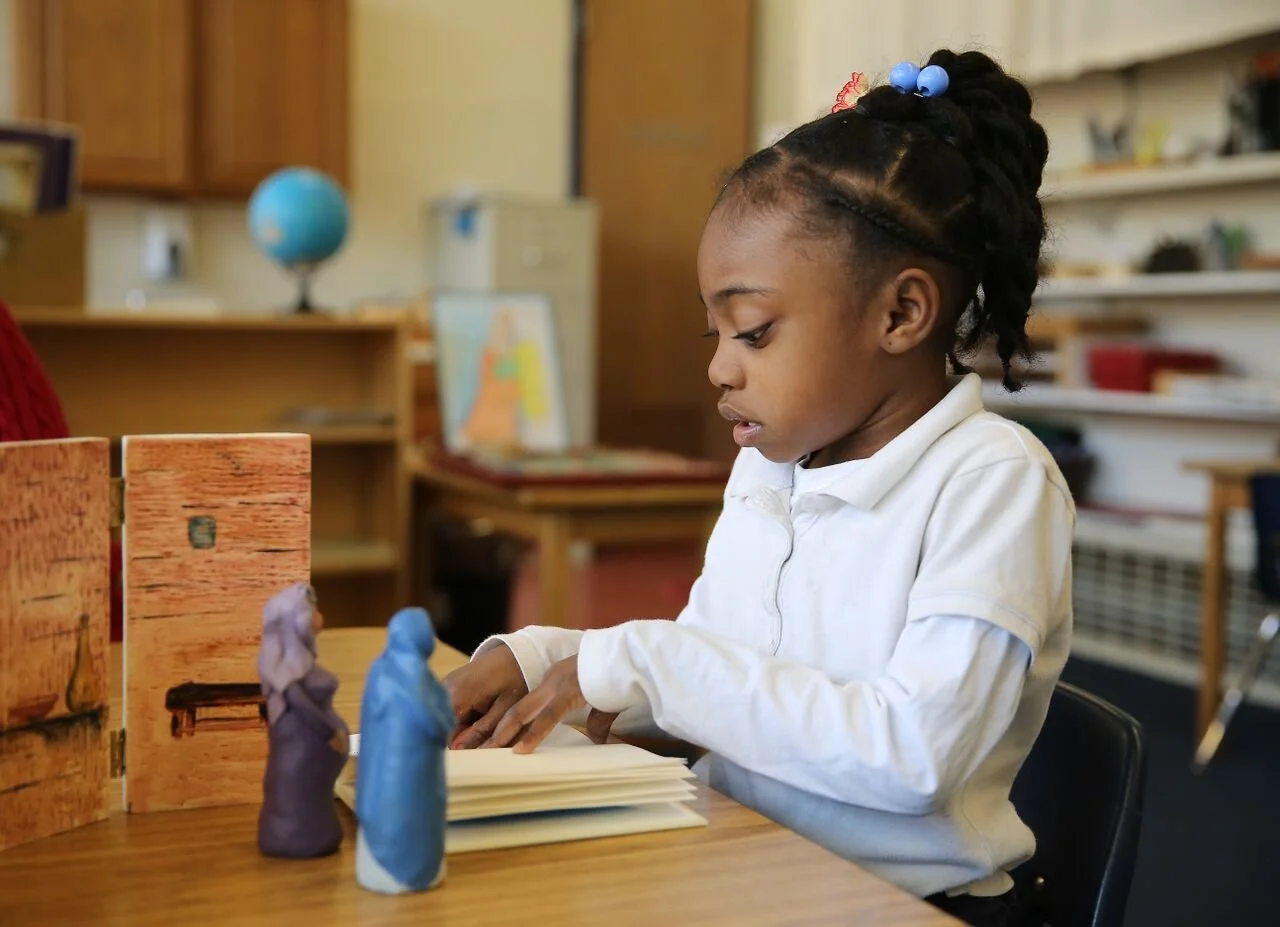 Young girl reading a book at a table with clay figures in a classroom setting.