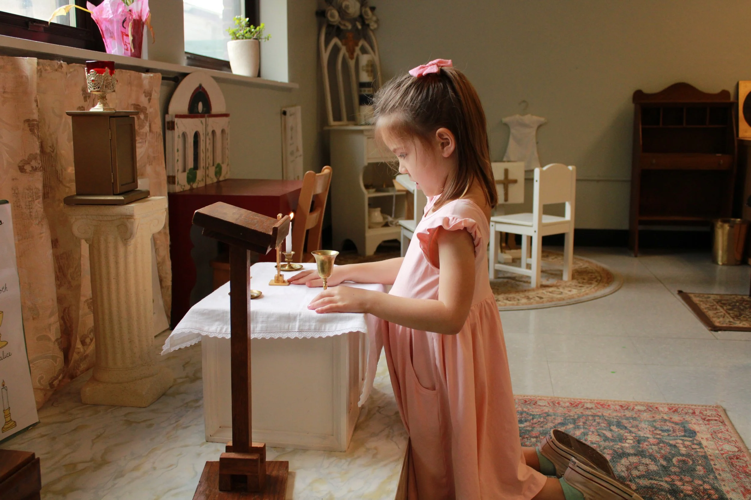 Young girl in a pink dress kneeling at a small altar with religious items, including a chalice and candles, in a classroom setting.