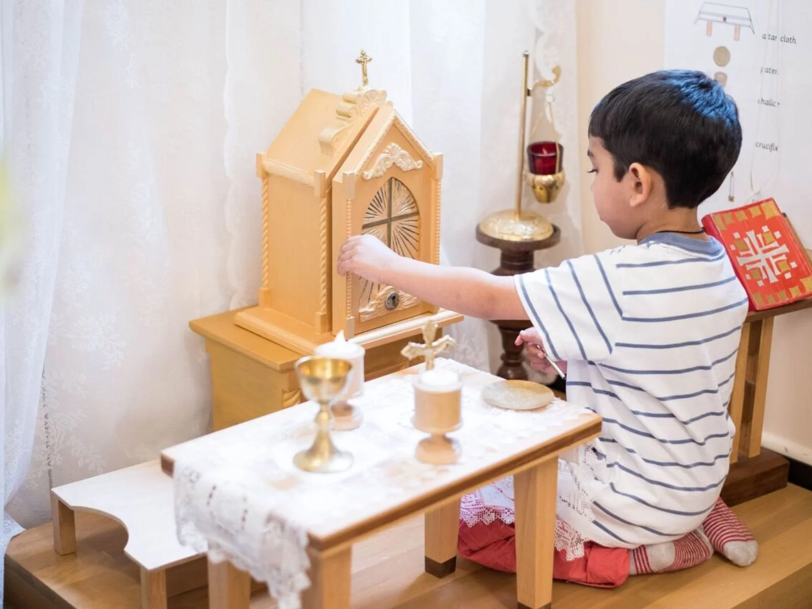 Young boy sitting on the floor, playing with a small wooden altar setup, including a tabernacle and miniature religious items, on a table covered with lace. He is wearing a striped shirt and red pants.