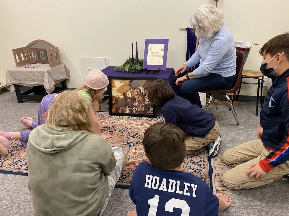 Group of children and a teacher observing a religious scene with nativity imagery displayed on a board, in a classroom setting.