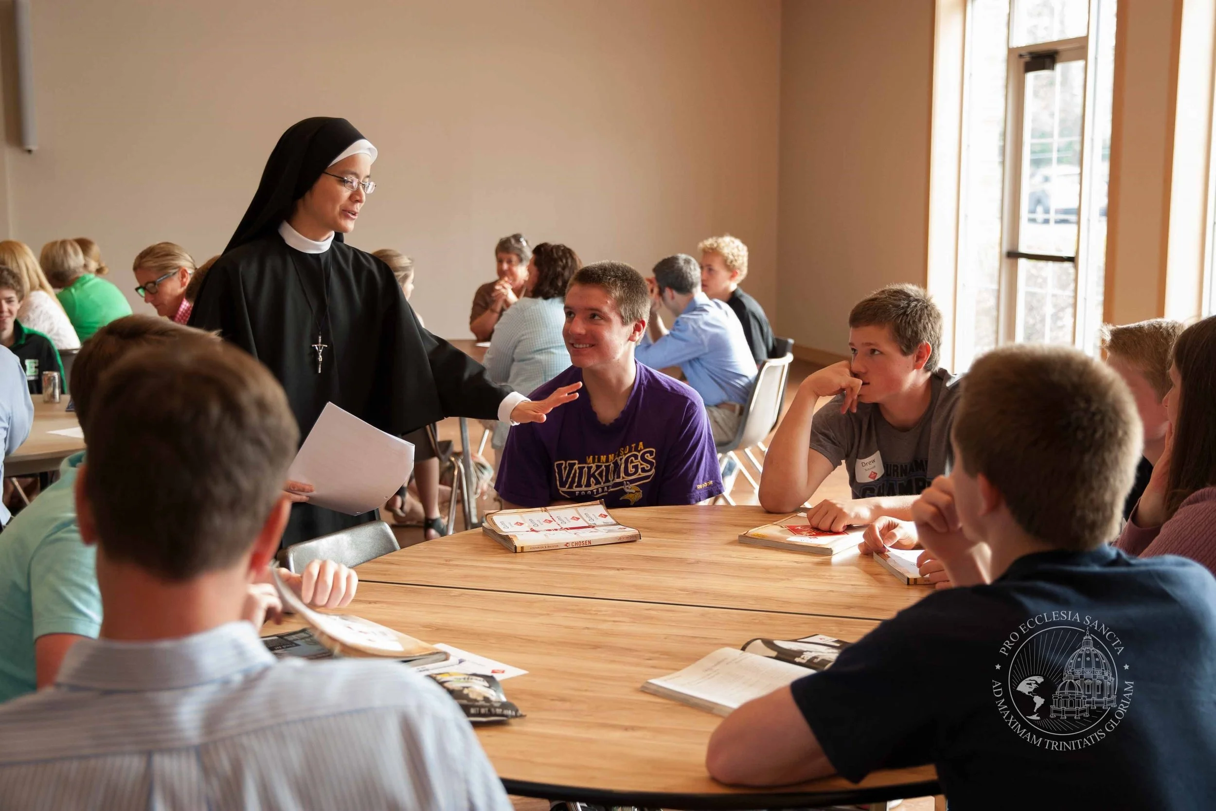 A nun wearing a habit is speaking to a group of teenagers seated around a table in a well-lit room. The students have books and notepads on the table.