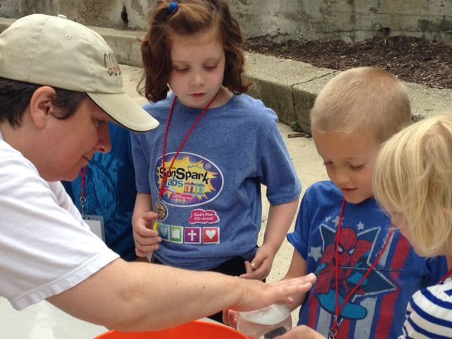 Children engaging in a science experiment led by an adult wearing a cap. They are touching ice floating in a bowl of water. The children are wearing name tags and event T-shirts.