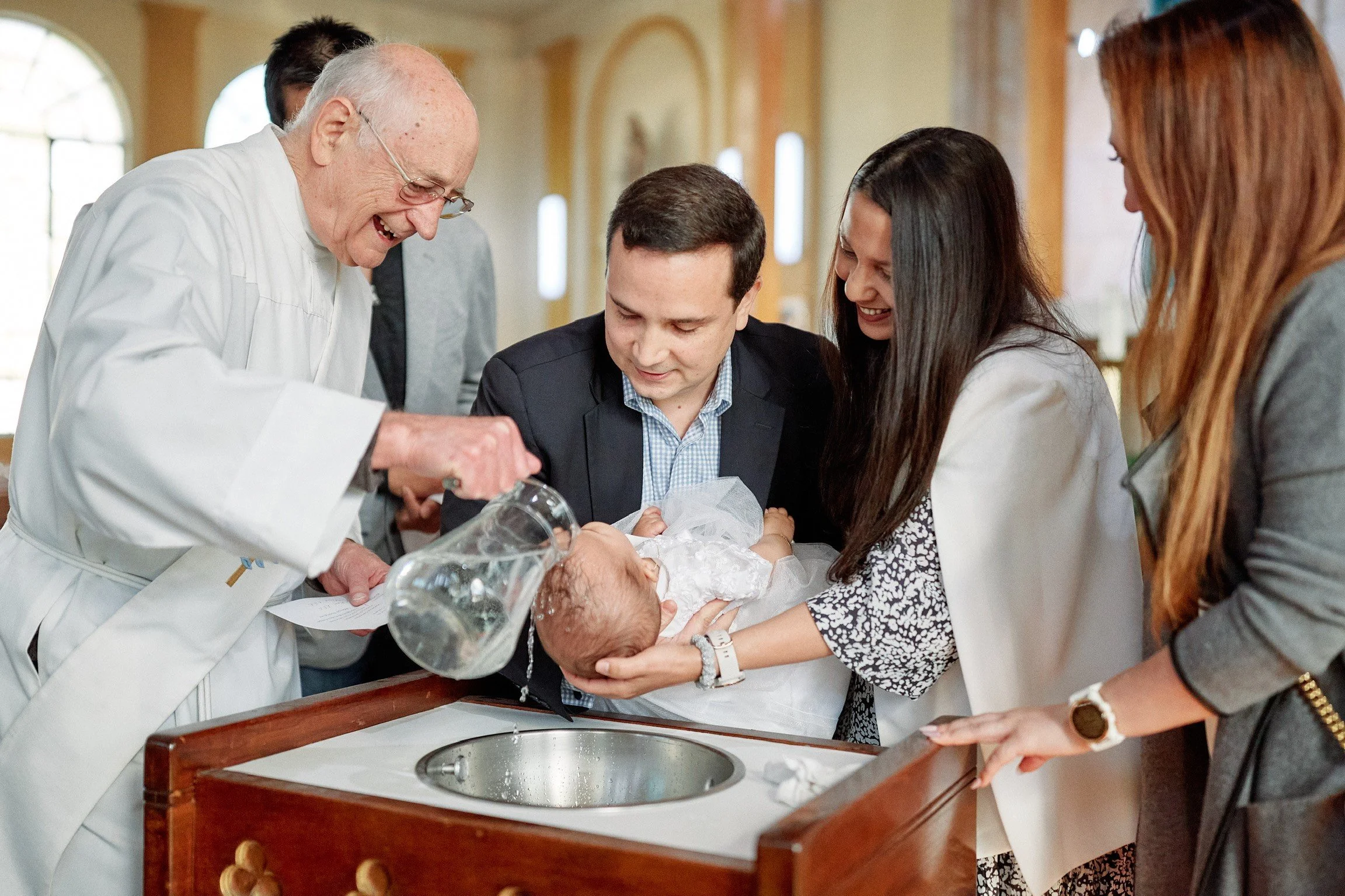 A baby being baptized by a priest, surrounded by family members, in a church setting.
