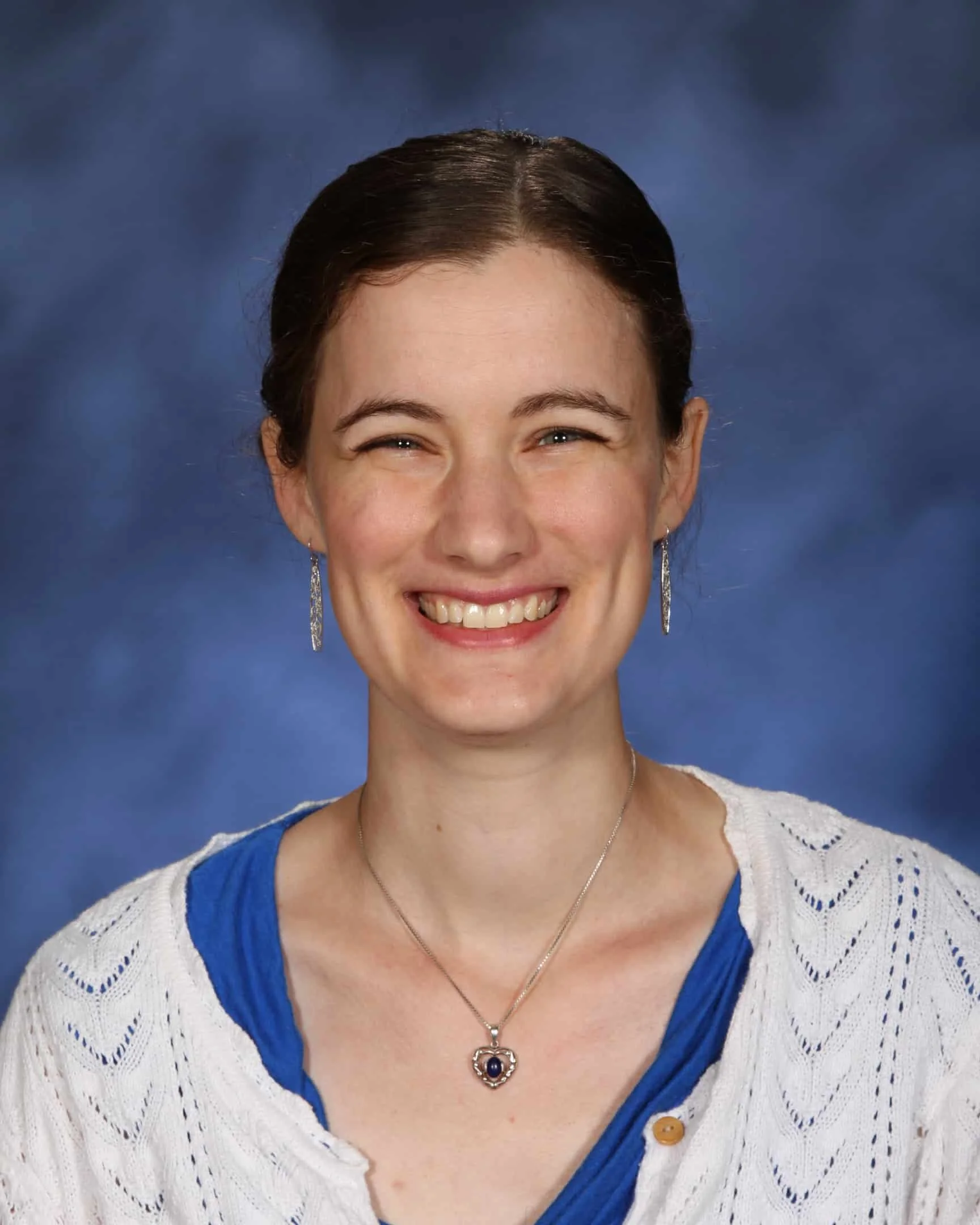 Smiling person wearing a white cardigan, blue top, heart-shaped pendant, and earrings, against a blue background.