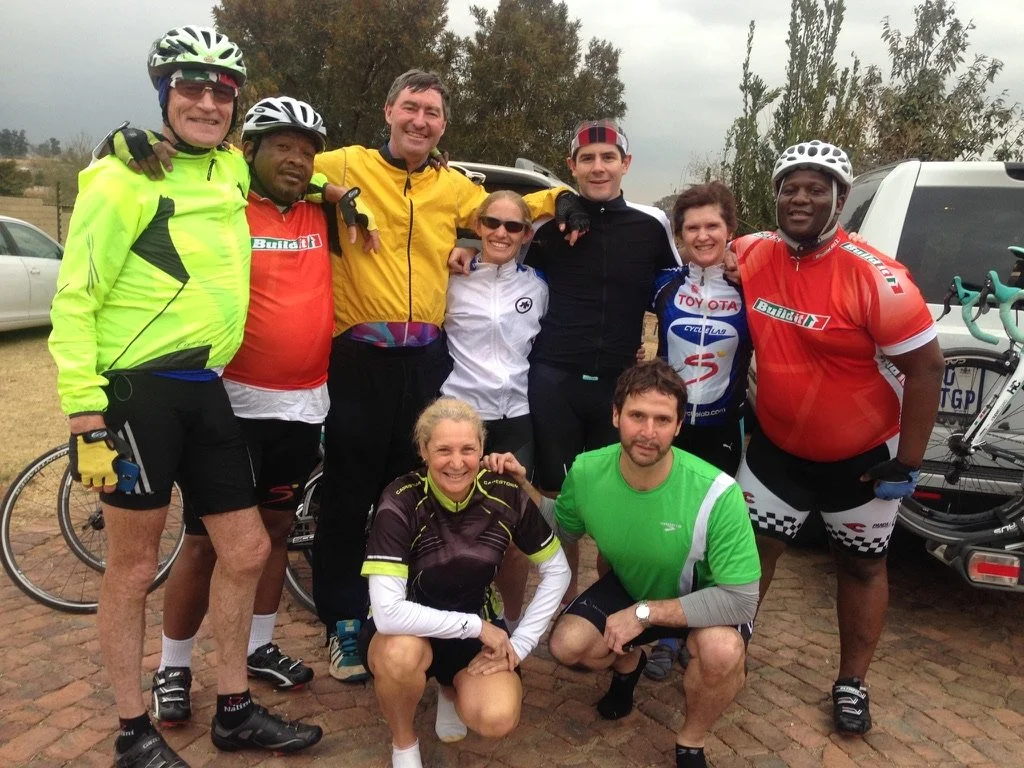 Group of nine cyclists posing outdoors on a cloudy day, with bikes and a vehicle in the background.
