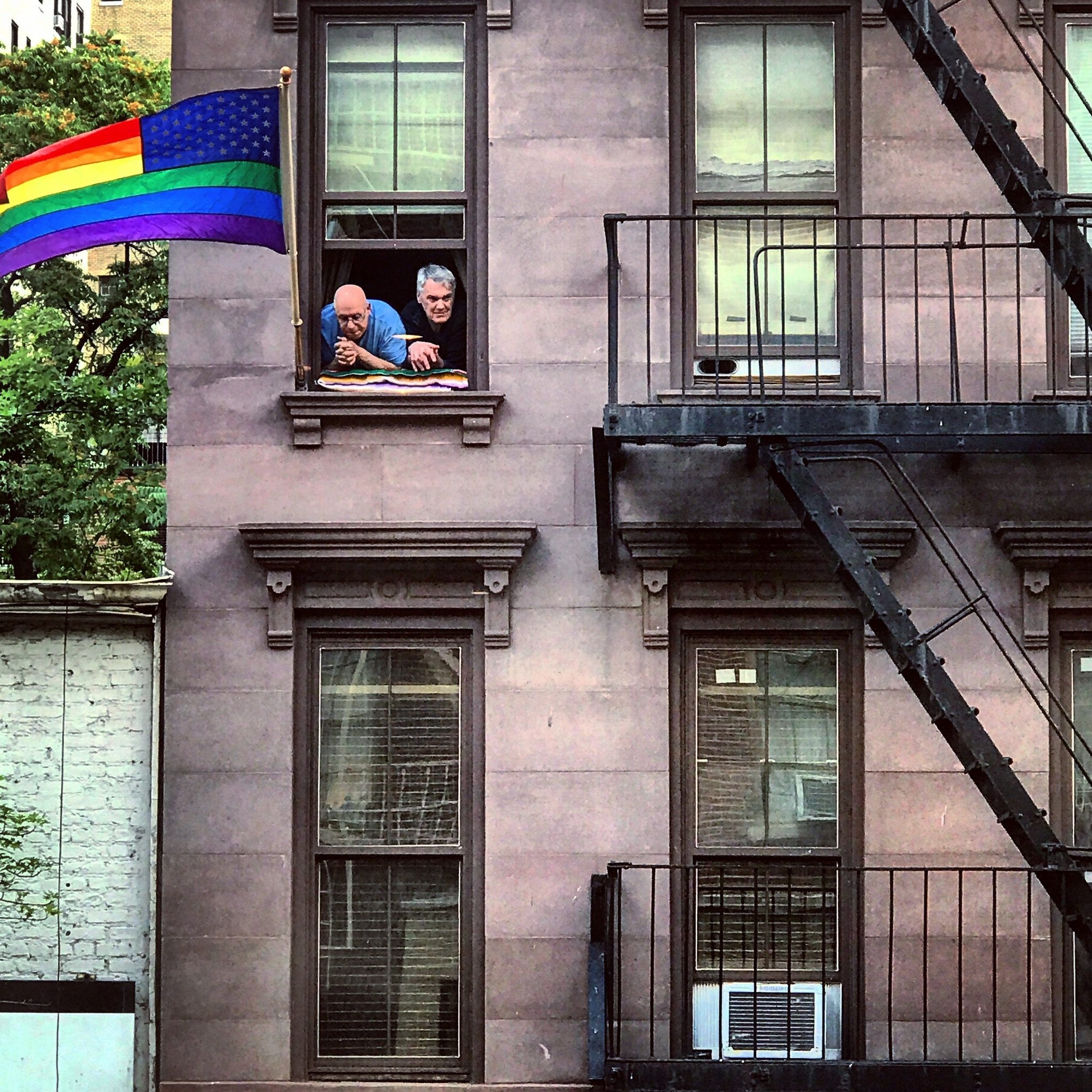 Two men looking out an apartment window with a pride flag, representing LGBTQ+ identity and affirming therapy support in NYC