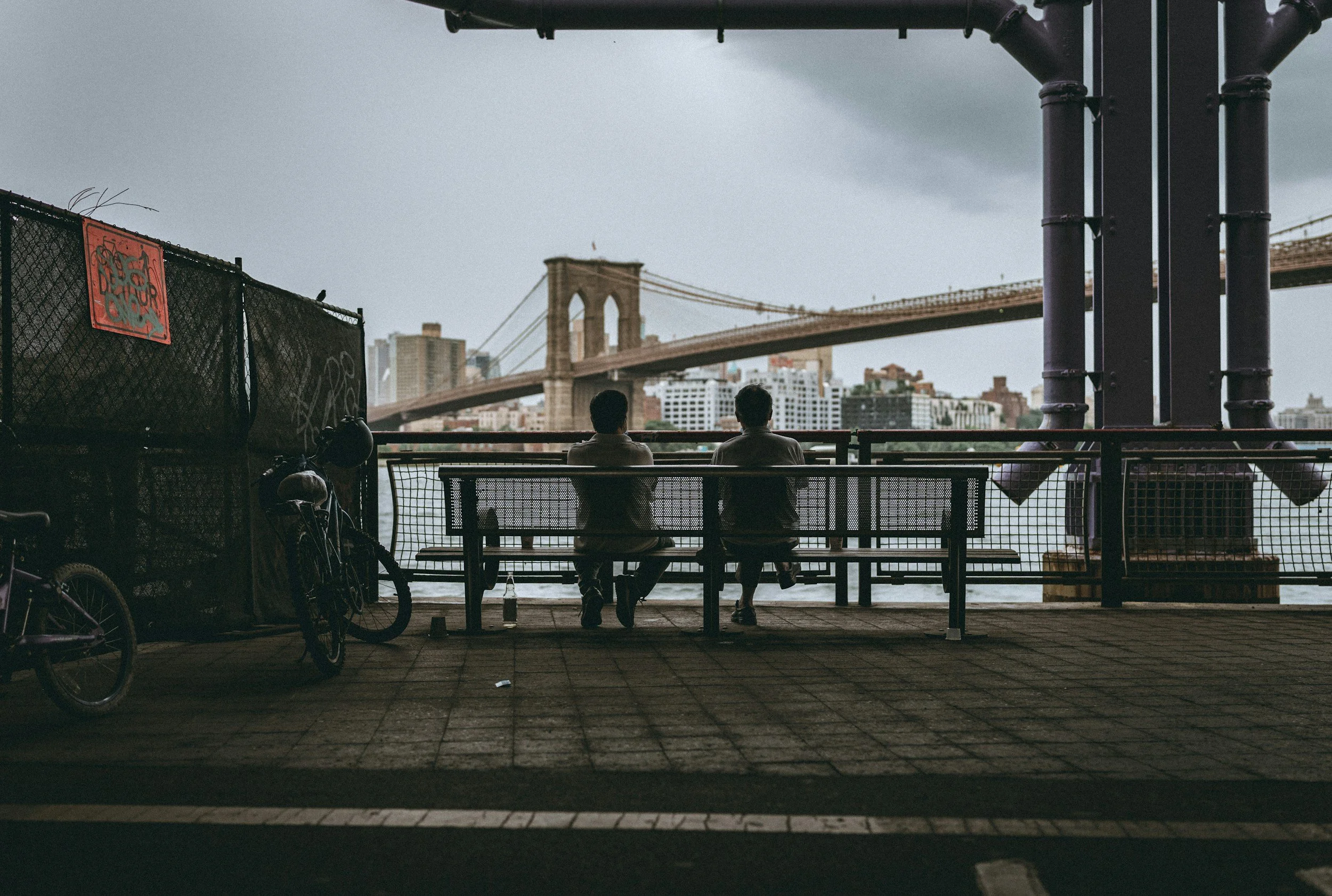 Two people sitting on a bench overlooking a bridge in New York City