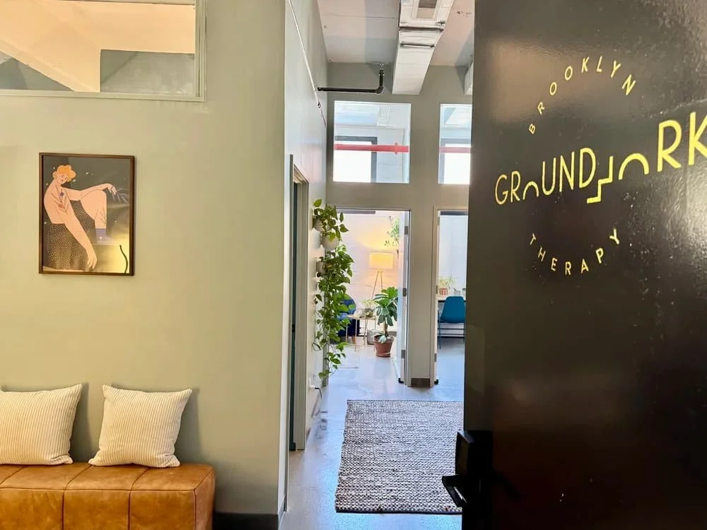 Interior of a therapy center named Brooklyn Groundjork Therapy, with a black wall featuring the center's yellow logo, a brown sofa with two cream pillows, a green wall with a framed artwork of a woman, and a corridor with potted plants leading to a bright room with windows.