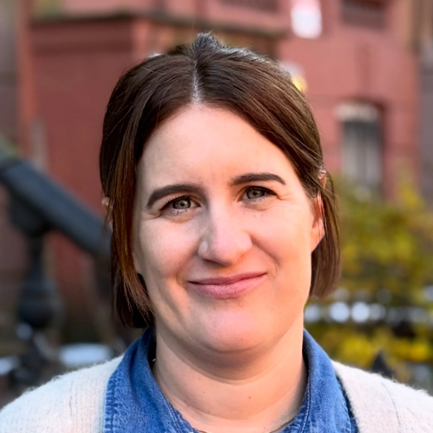 A woman with long, straight brown hair smiling outdoors on a city street with trees and brick buildings in the background.