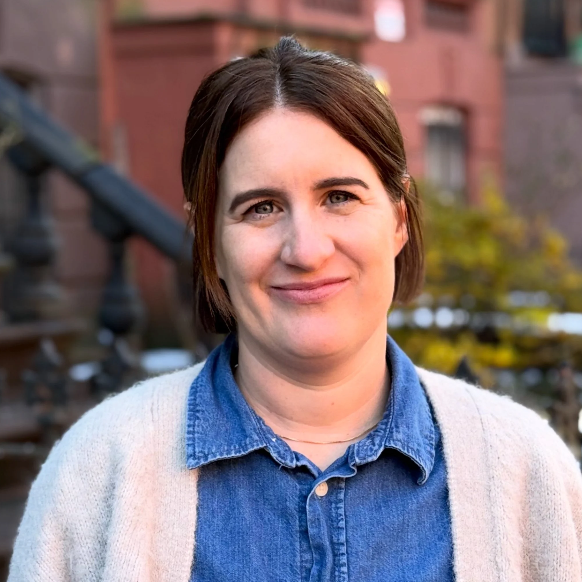 A woman with short brown hair wearing a blue denim shirt and a beige cardigan, standing outdoors in front of brick buildings and trees.