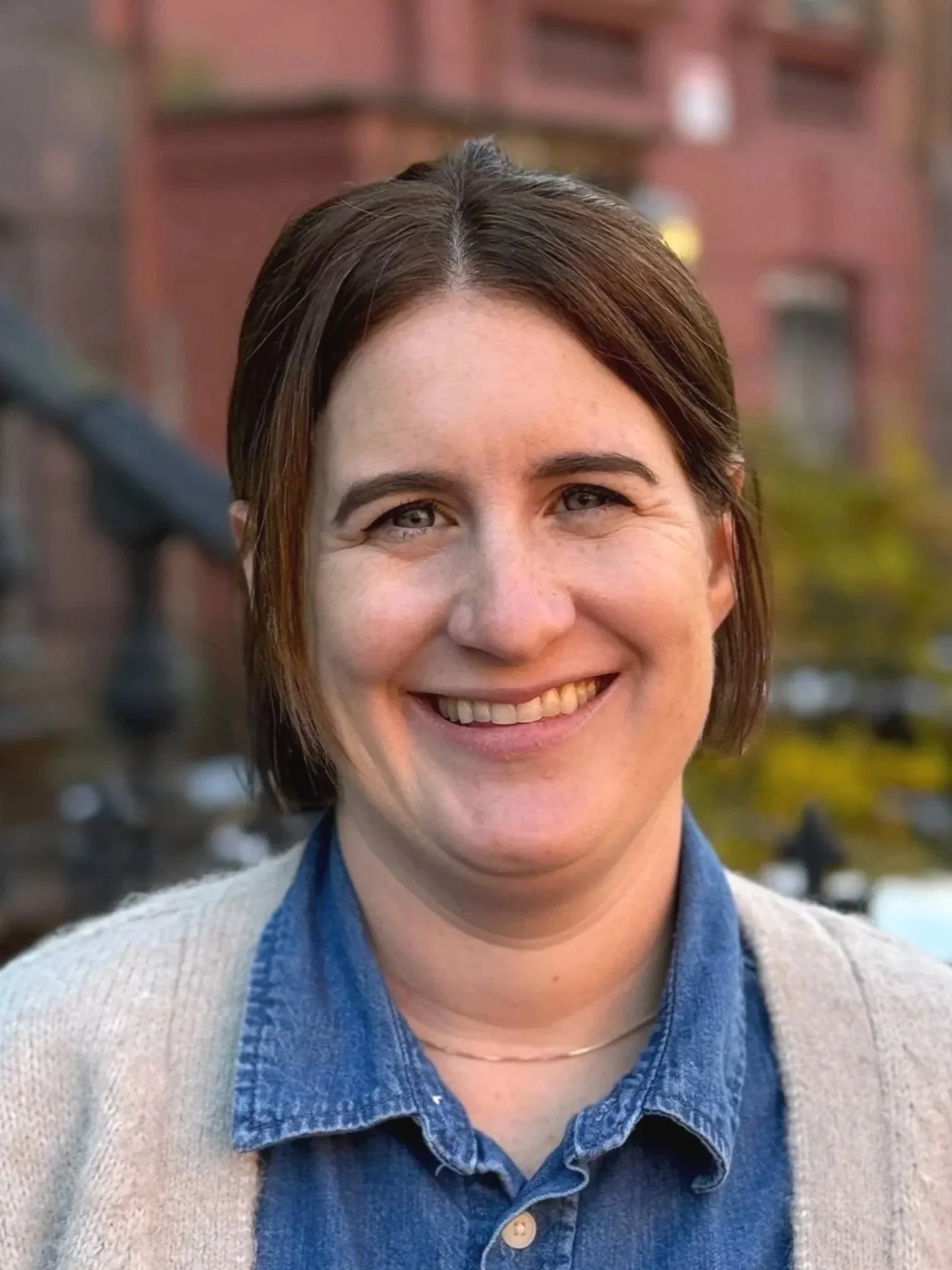 A woman with long, straight brown hair smiling outdoors on a city street with trees and brick buildings in the background.