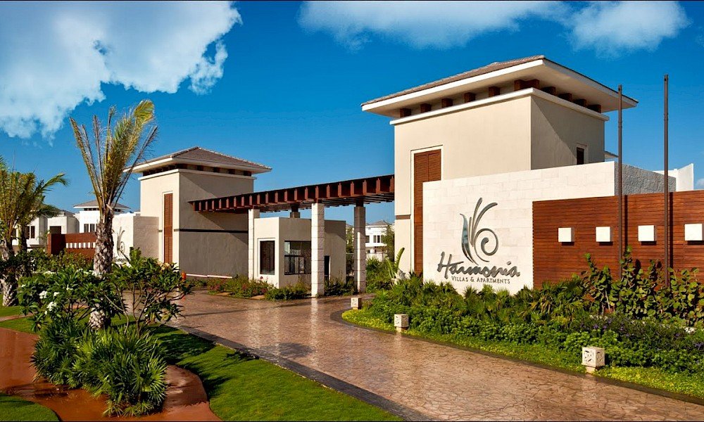 Entrance of a residential complex with modern villas, palm trees, green landscaping, and a white wall with the name 'Hacienda Villas & Apartments' visible.