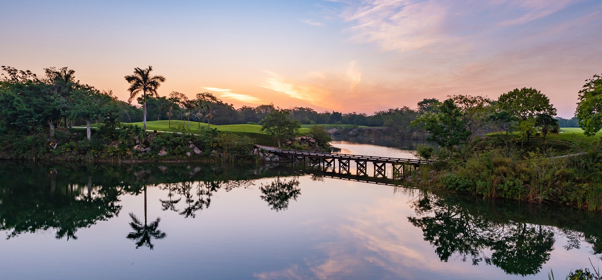Sunset over a lush green area with palm trees, a wooden bridge over a calm body of water, and a sky with pink, purple, and orange clouds reflected in the water.