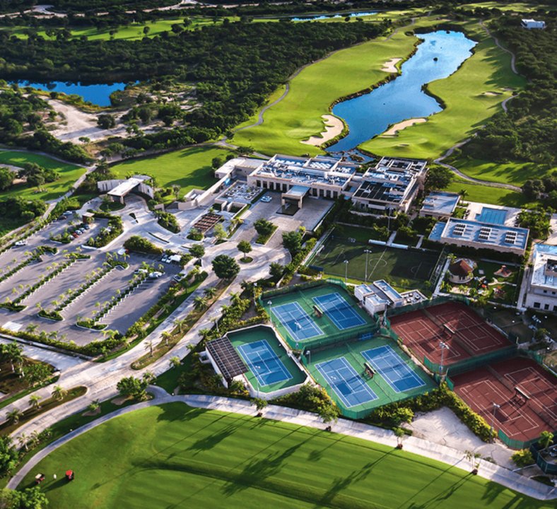 Aerial view of a sports complex with multiple tennis courts, a football field, an academic building, and surrounding greenery and water bodies.