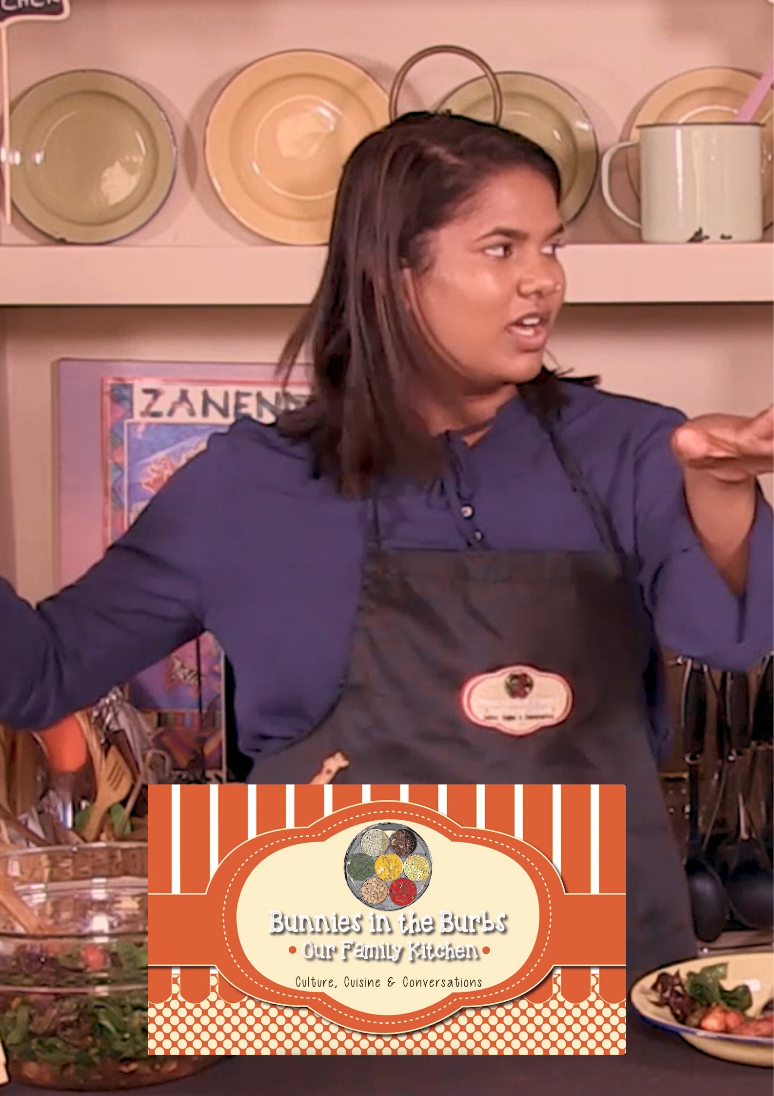 A woman with dark hair wearing a black apron and blue shirt gesturing with her arms in a kitchen. Shelves with plates, bowls, and kitchenware are behind her. A sign on the wall reads "Zanena" and another graphic footed board displays the text "Bunnies in the Burbs Our Family Kitchen - Culture, Cuisine & Conversations."