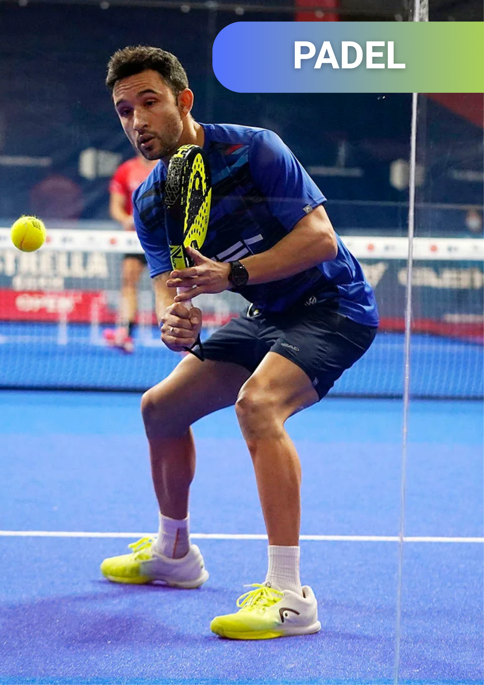 A man playing padel on an indoor court, holding a yellow padel racket, with a yellow tennis ball in mid-air nearby, wearing a blue shirt, black shorts, white socks, and yellow sports shoes.