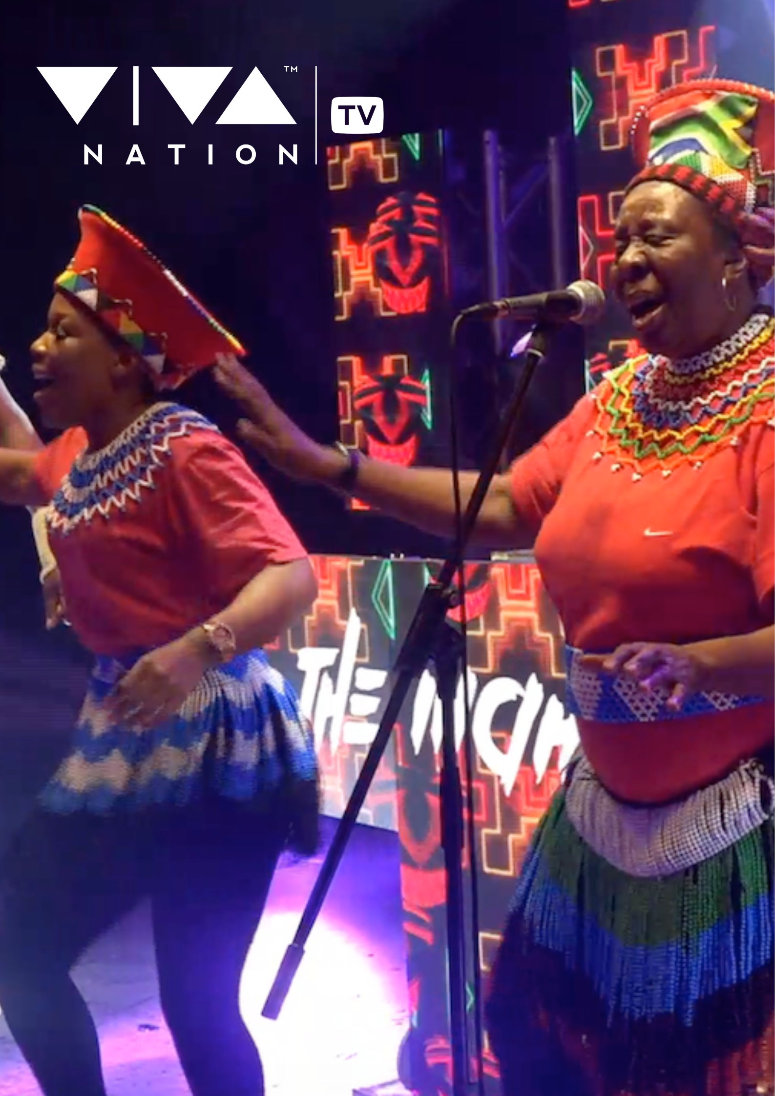 Two women dressed in traditional African attire singing and dancing on stage with colorful neon lights and tribal motifs in the background.