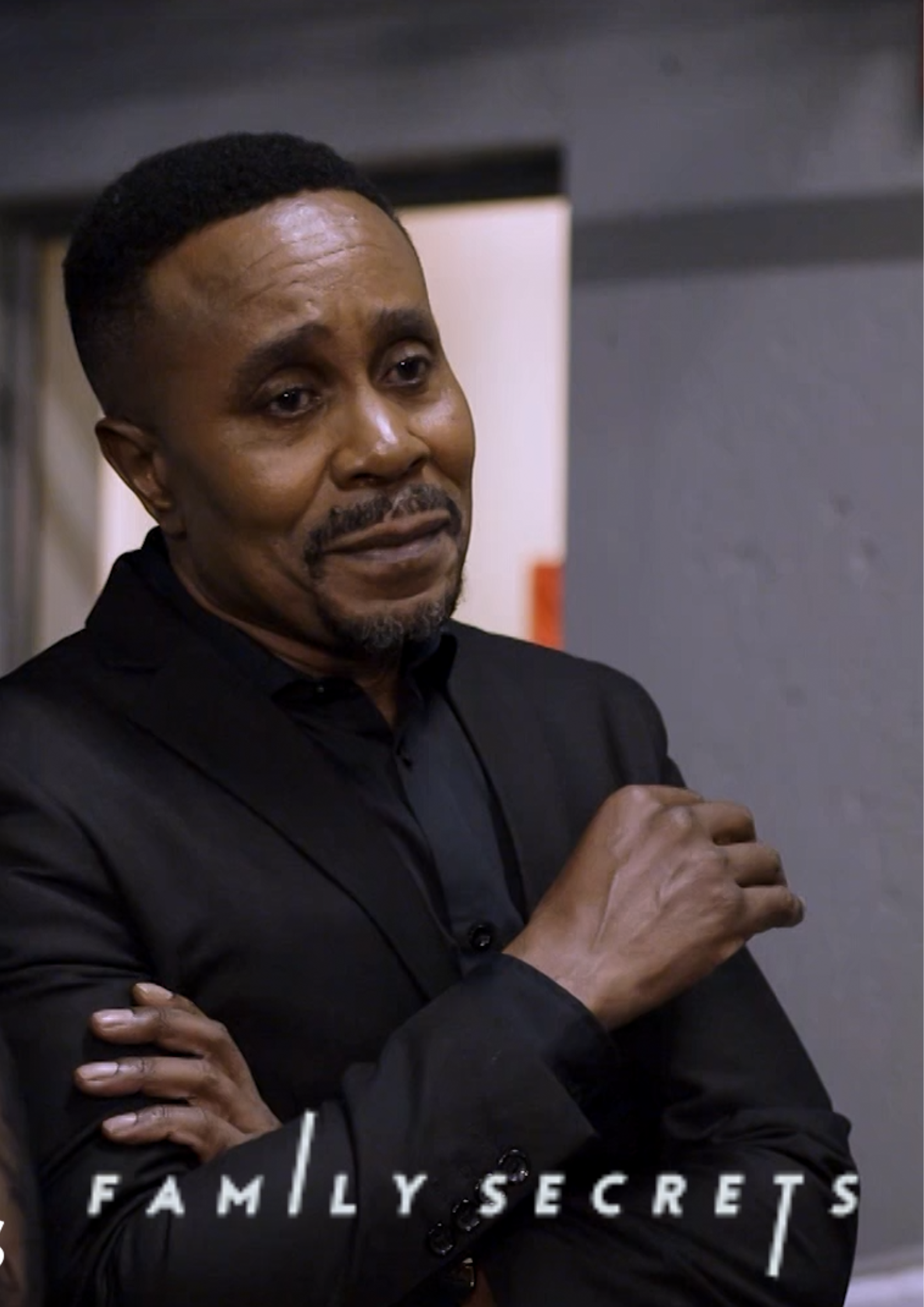 A man in a black suit with a sad or contemplative expression standing with his arms crossed. The background is blurred, and there is a logo that reads "FAMILY SECRETS" at the bottom.