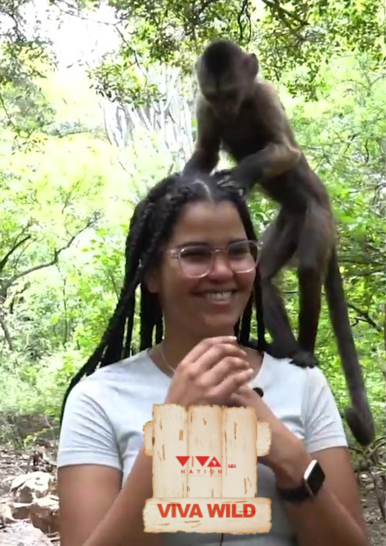 A smiling woman with glasses and braided hair in a forest, with a monkey on her shoulders and another monkey grabbing her hair.
