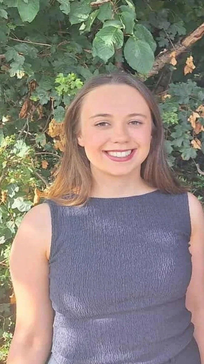 A young woman with long, light brown hair smiling outdoors in front of green leafy bushes, wearing a sleeveless dark blue top.