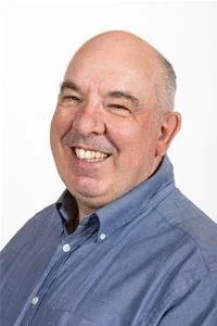 Smiling middle-aged man with a bald head wearing a blue button-up shirt against a white background.