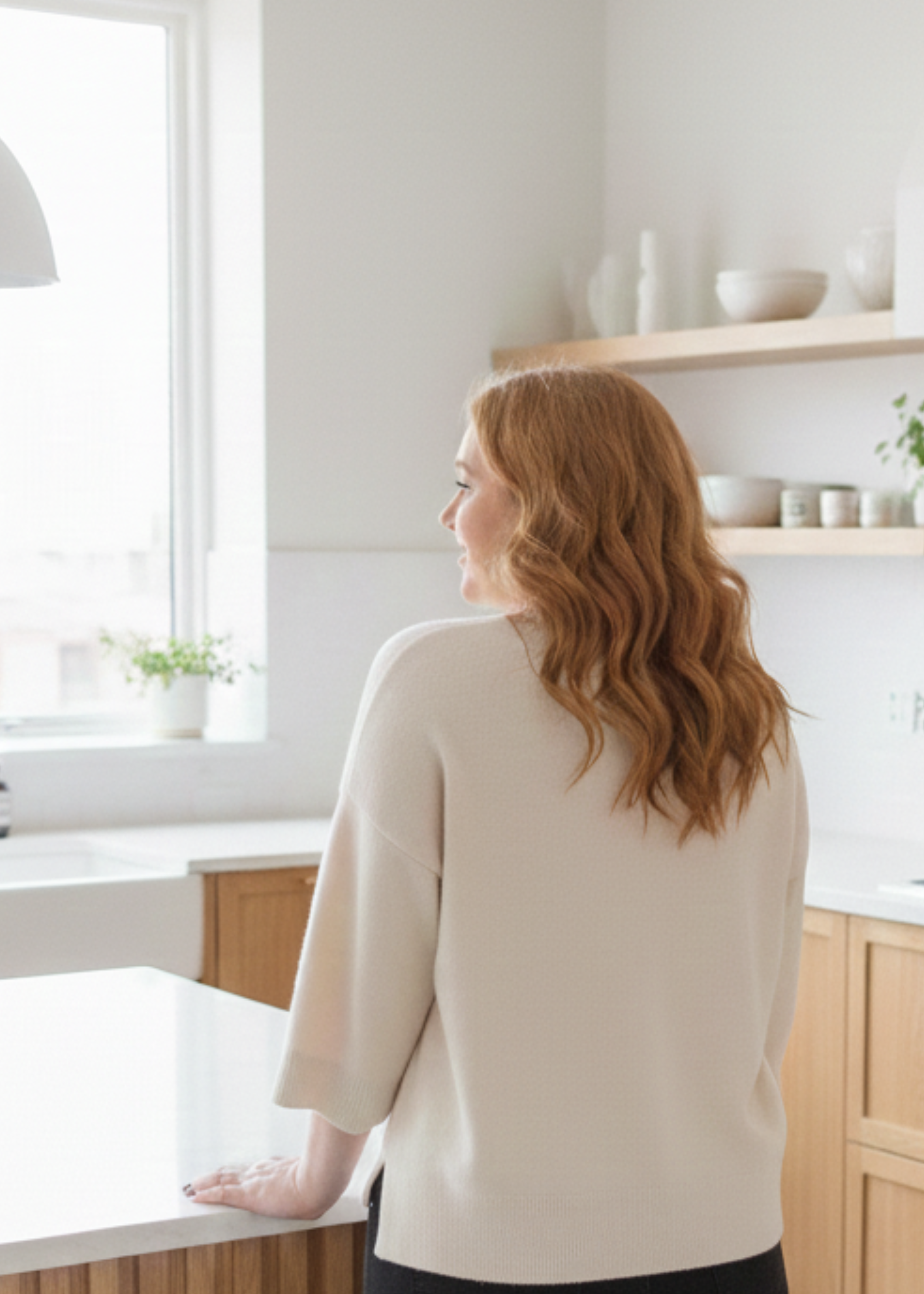 A woman with red hair and styled waves standing in a modern, bright kitchen with her back to the camera, looking out the window.