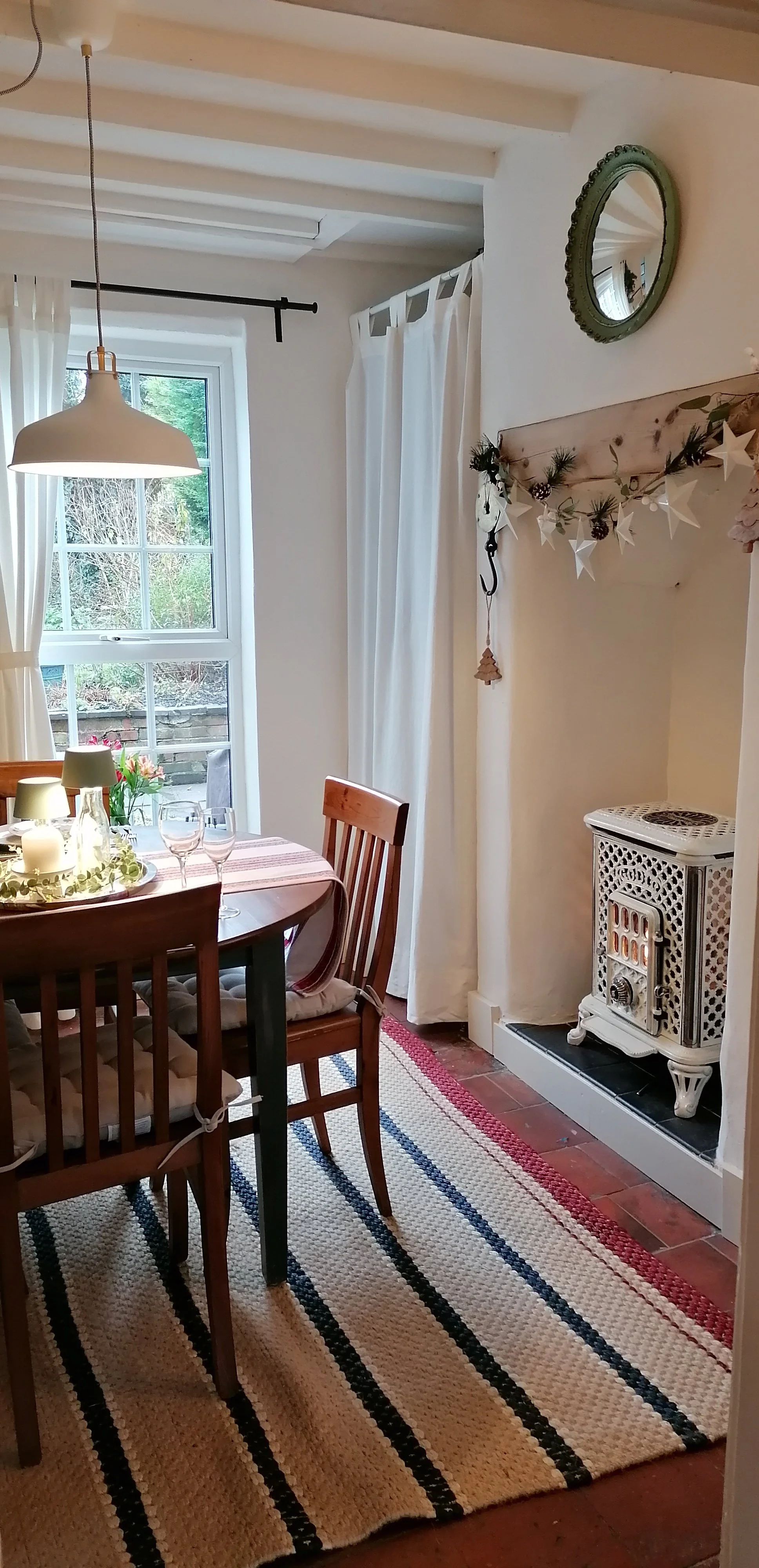 Cozy dining room with wooden table, chairs, a striped rug, a window with white curtains, and a white heater in the corner decorated with a holiday banner.