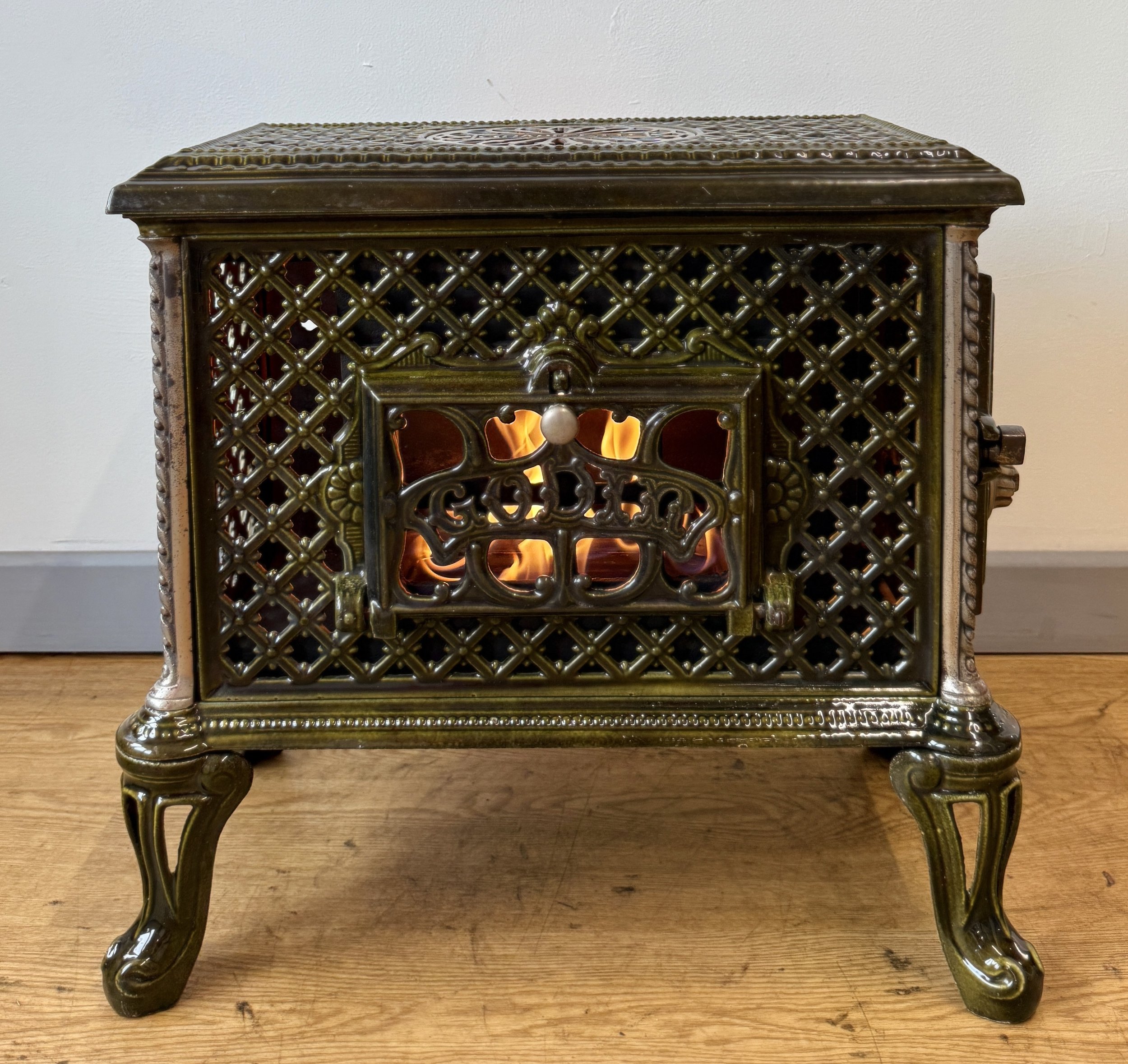 An antique cast iron decorative heater with flames inside, sitting on a wooden floor against a white wall.