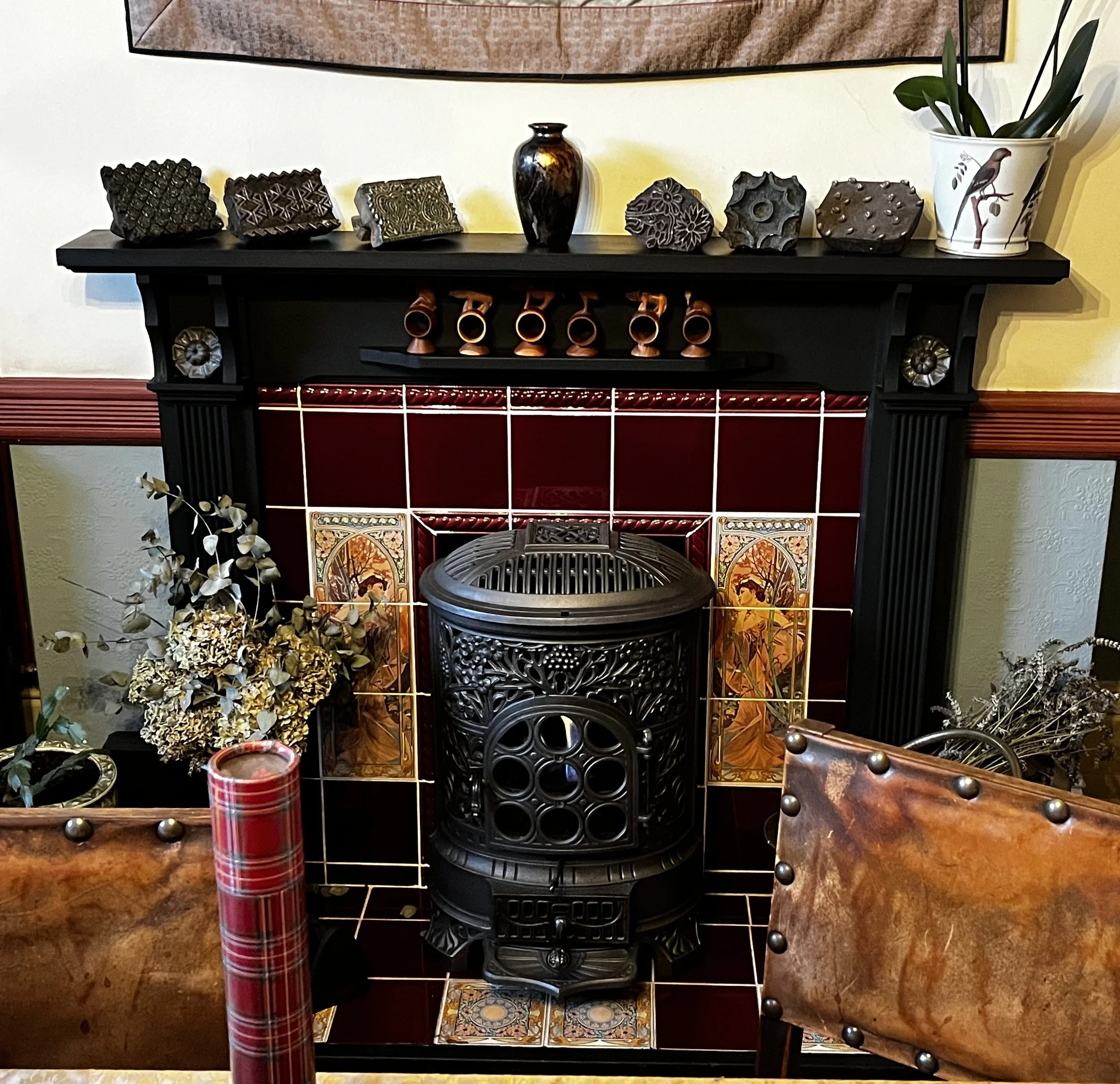 A room with a black fireplace mantel decorated with various art pieces and a potted plant, a vintage cast iron stove in front, and chairs with brown leather seats nearby.