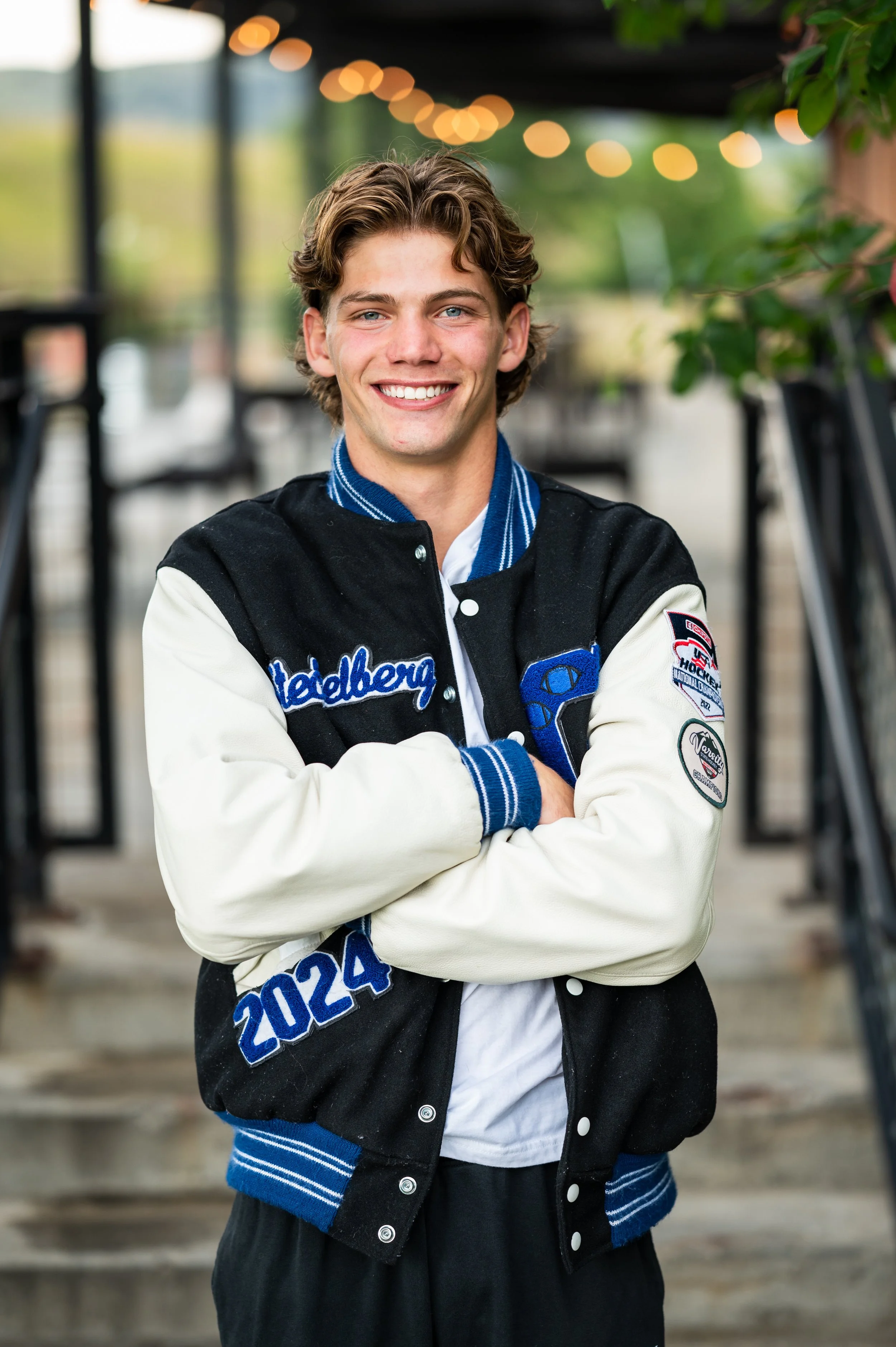 Young man with curly hair smiling, wearing a black and white varsity jacket with patches, standing outdoors with greenery and string lights in the background.