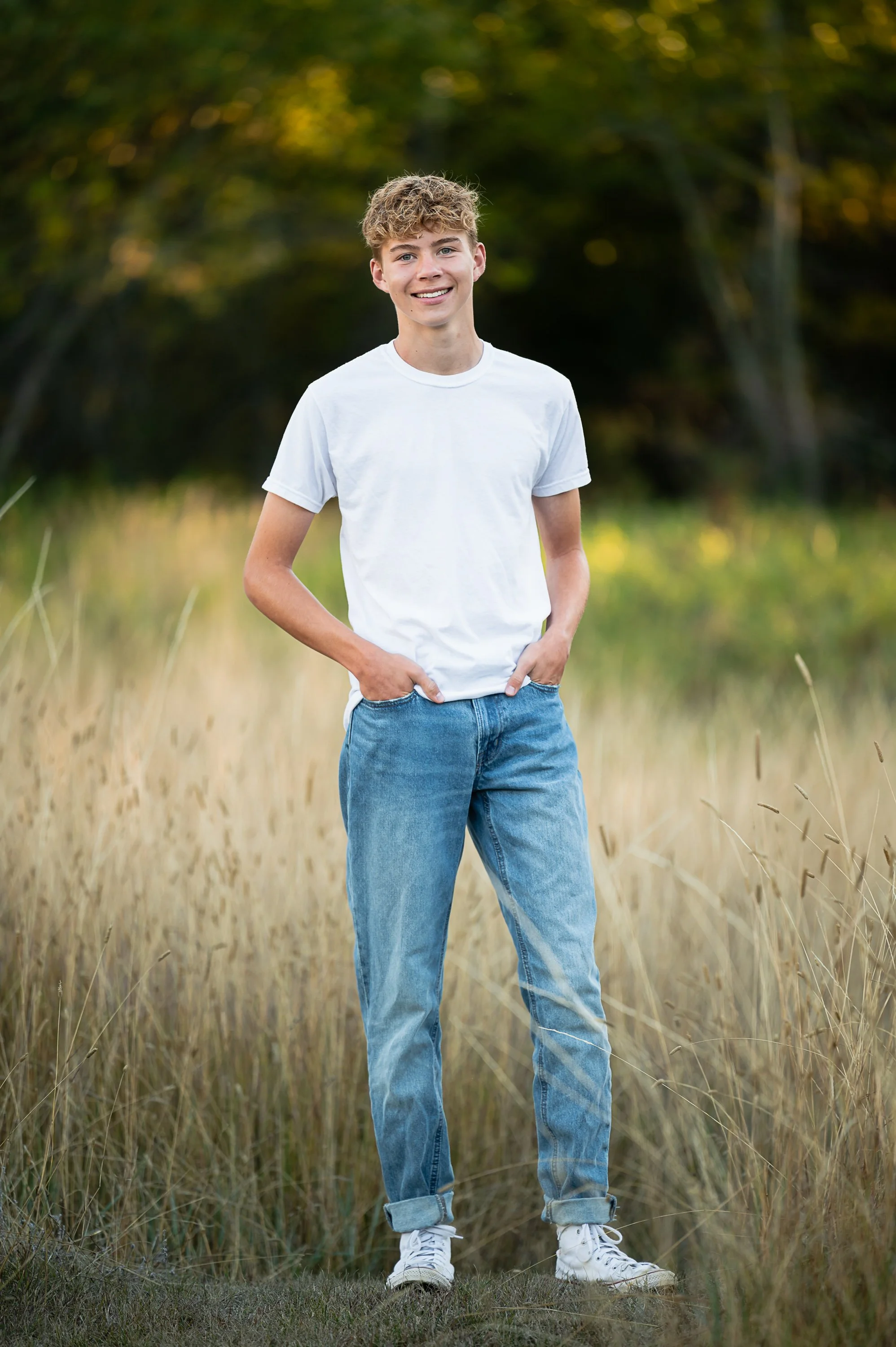 A young man stands outdoors in a grassy field with trees in the background, smiling and wearing a white t-shirt and light blue jeans.