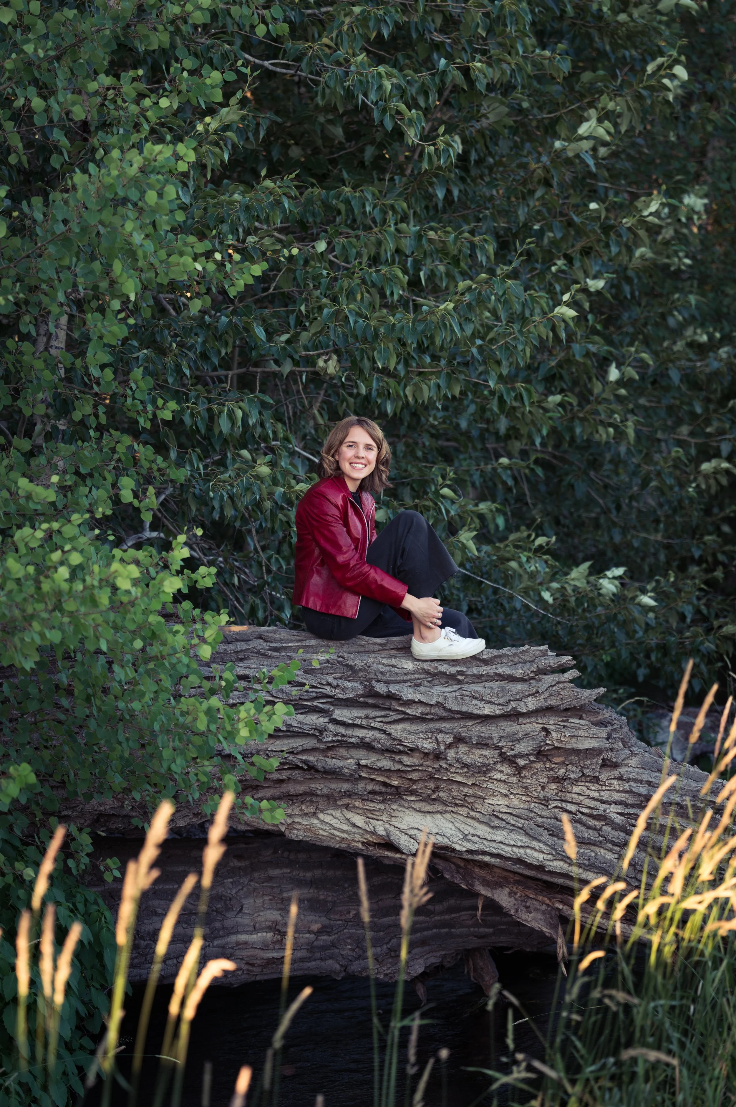 A young woman with shoulder-length brown hair, wearing a red jacket, black pants, and white shoes, sitting on a fallen tree trunk in a lush green forest, smiling at the camera.