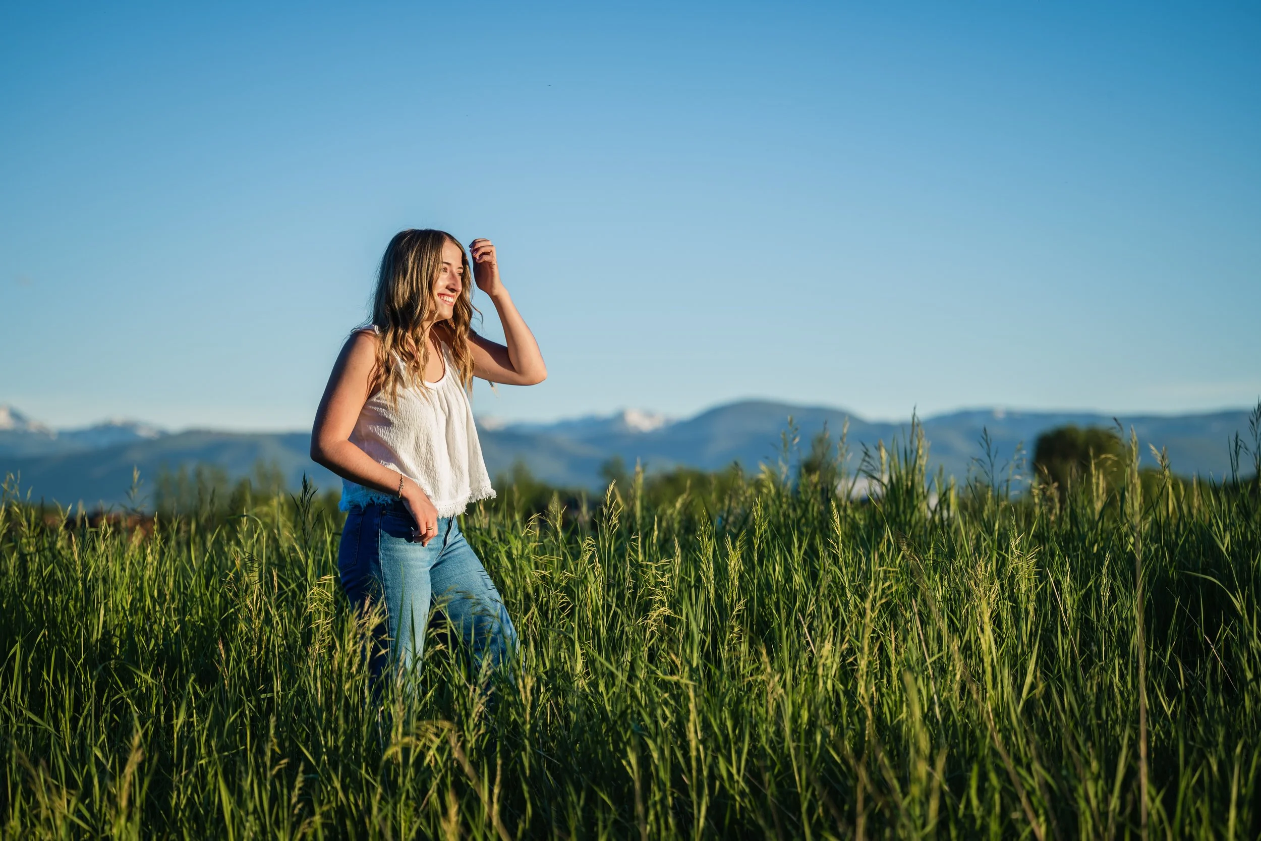 A young woman in a white top and jeans standing in a green field of tall grass, smiling and enjoying sunshine with mountains in the background.