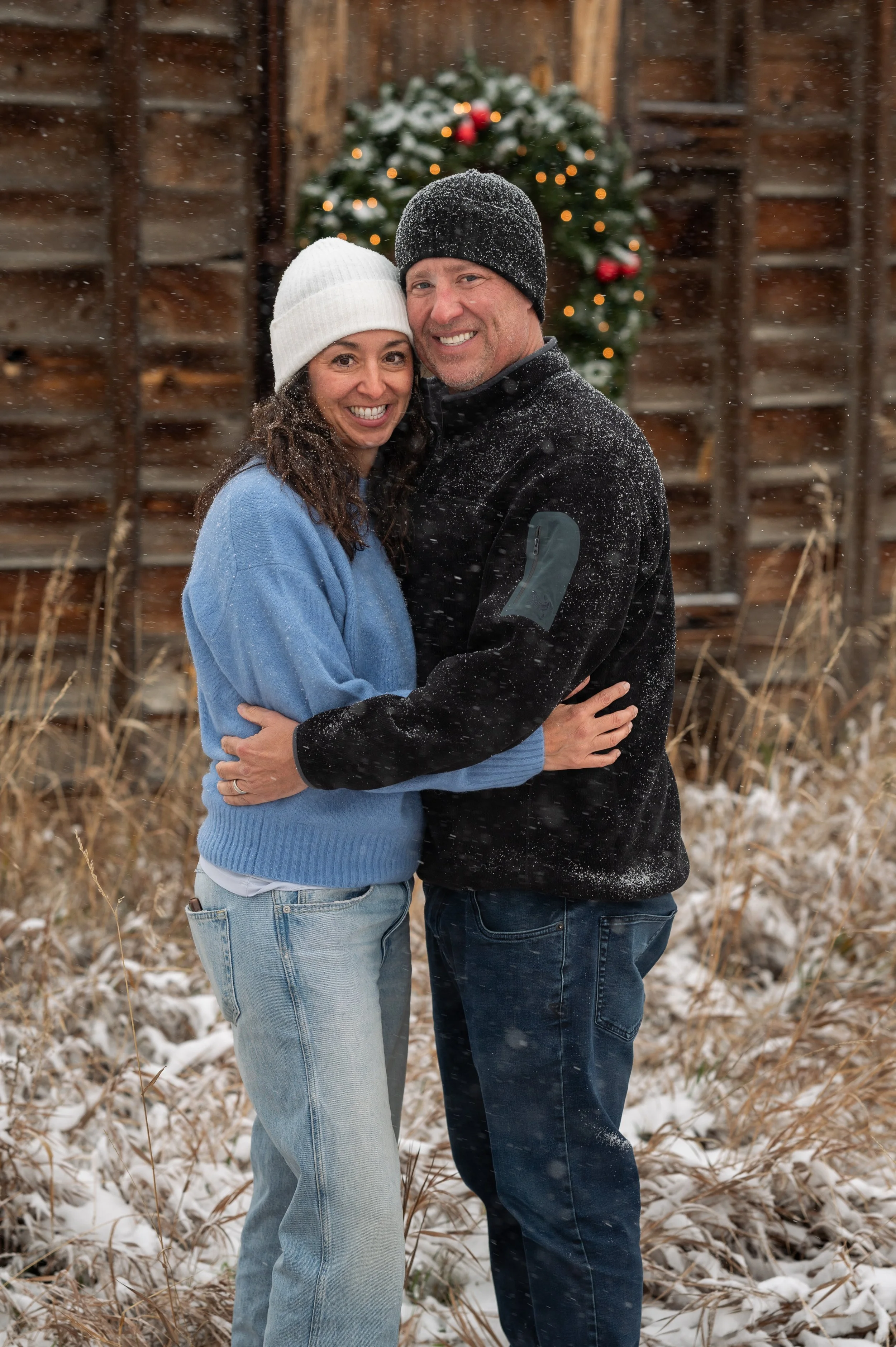 A smiling couple embraces outdoors in winter with snow falling, behind a Christmas wreath on a rustic wooden wall.