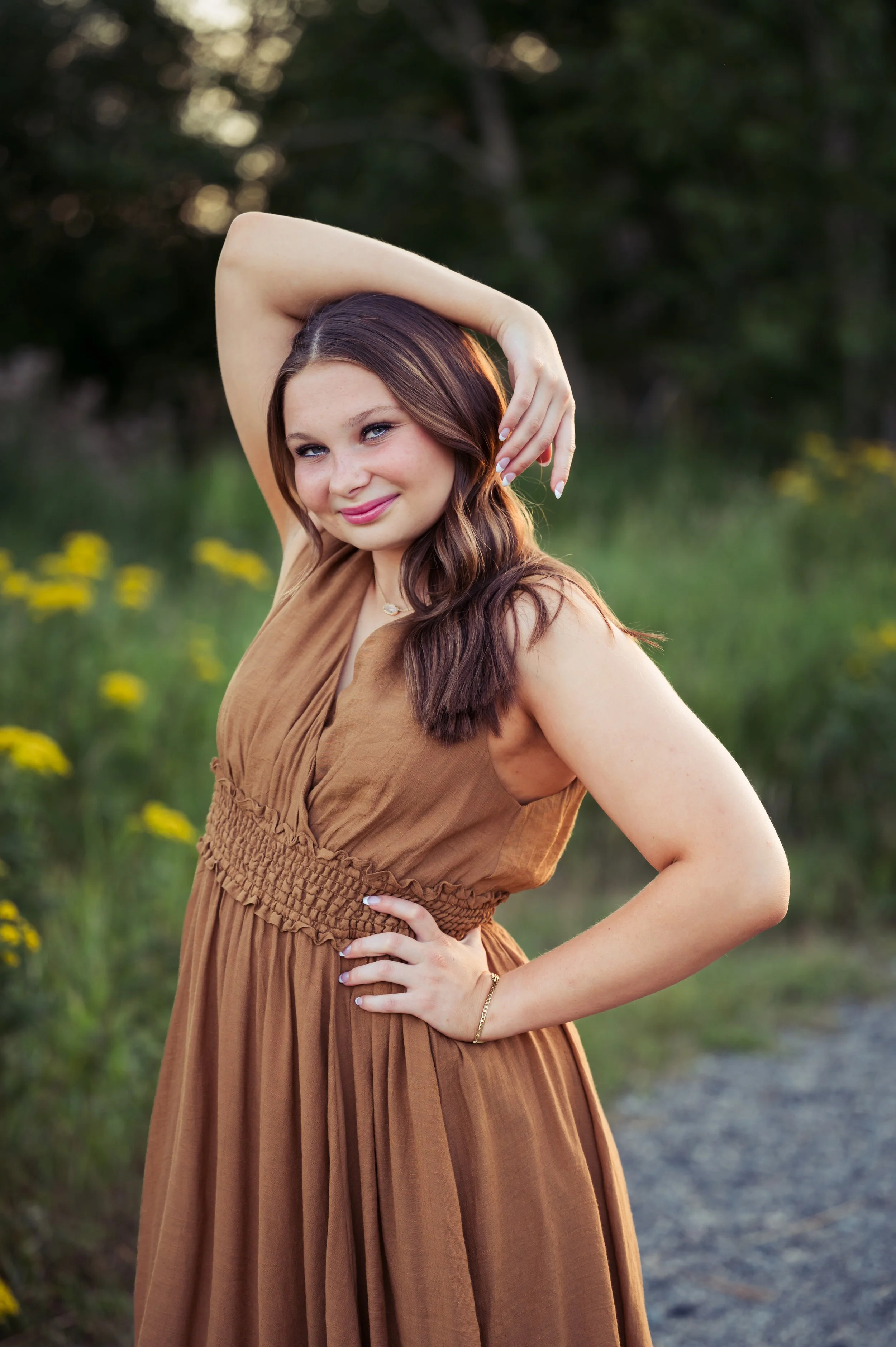 A young woman in a brown dress posing outdoors with hand on her hip, smiling at the camera in a natural setting with green grass and yellow flowers.