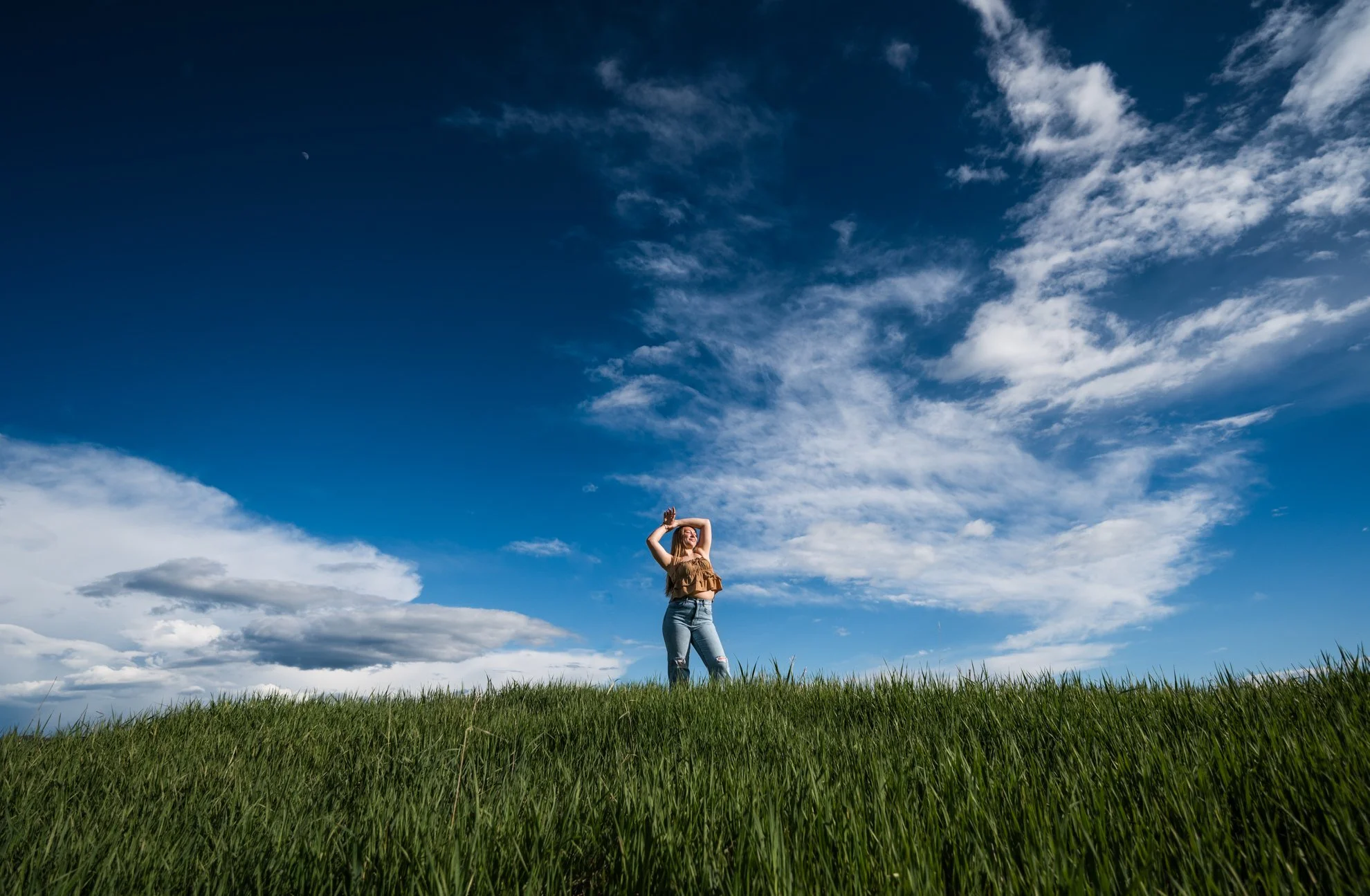 A woman standing in tall green grass under a blue sky with scattered clouds, holding her hands above her head and looking up.