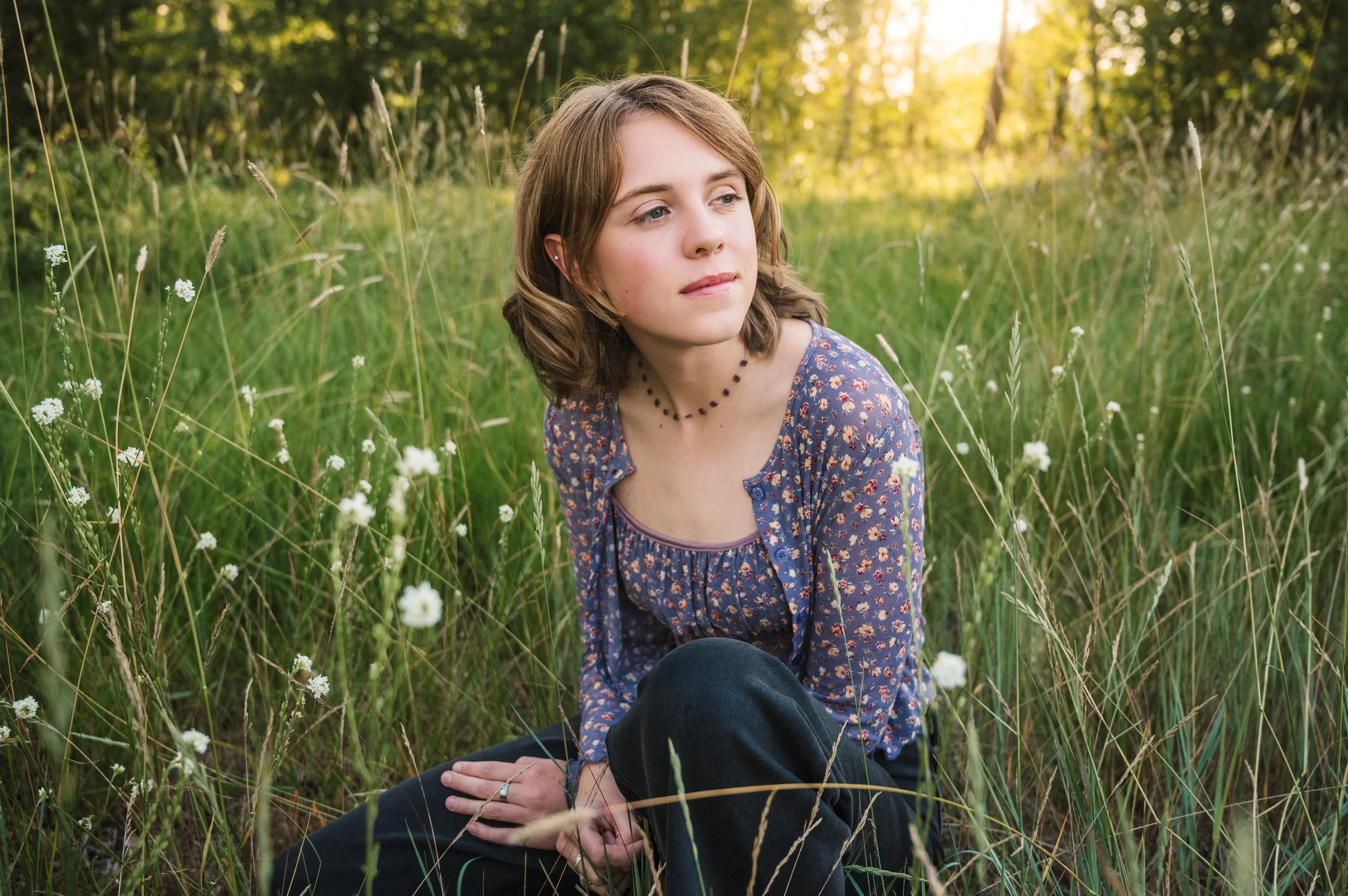 A young woman with light brown hair sits amidst tall grass and wildflowers in a field, looking thoughtfully into the distance during sunset.