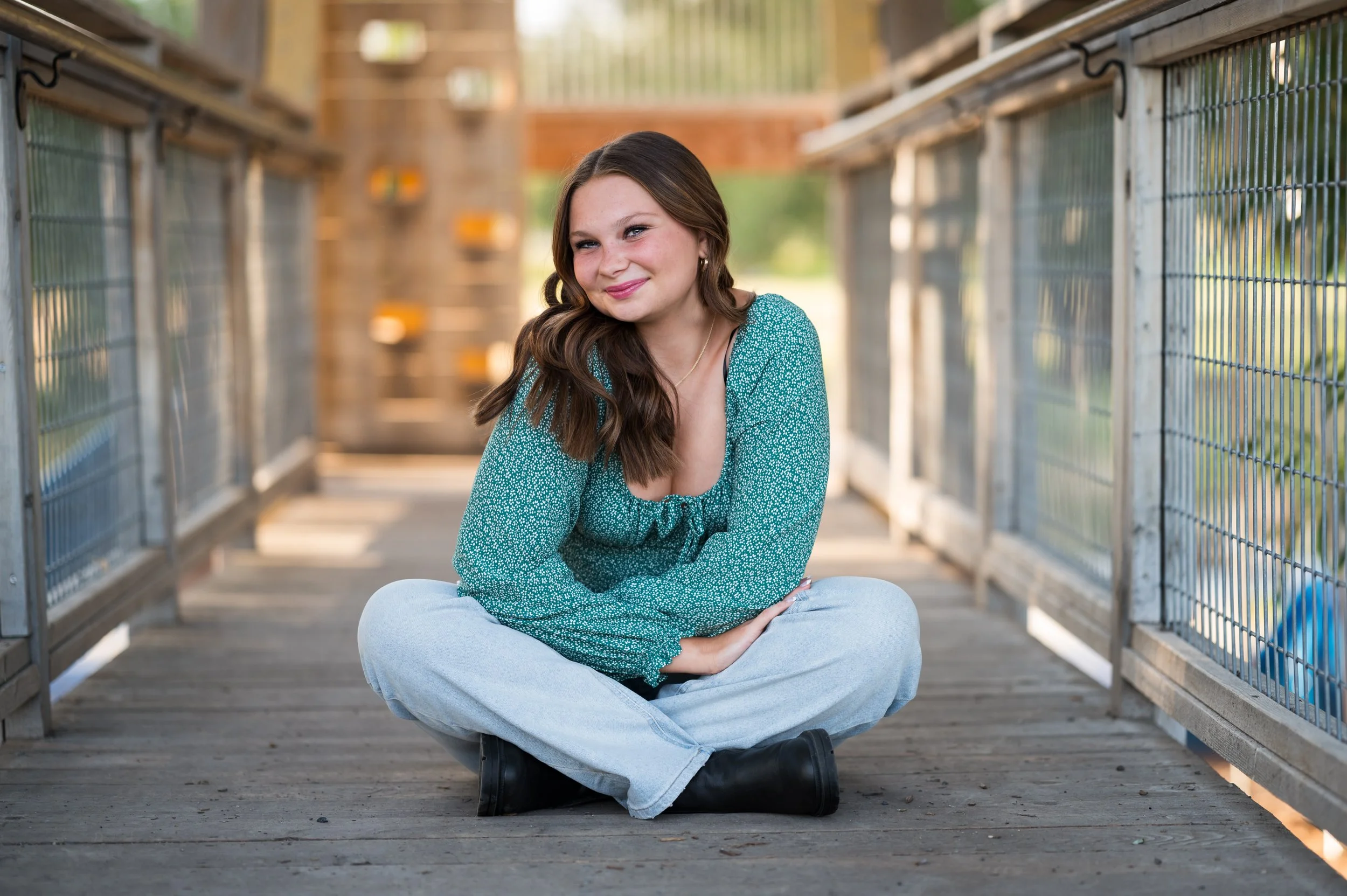 A young woman with long brown hair, wearing a turquoise blouse and light jeans, sitting cross-legged on a wooden bridge outdoors, smiling at the camera.