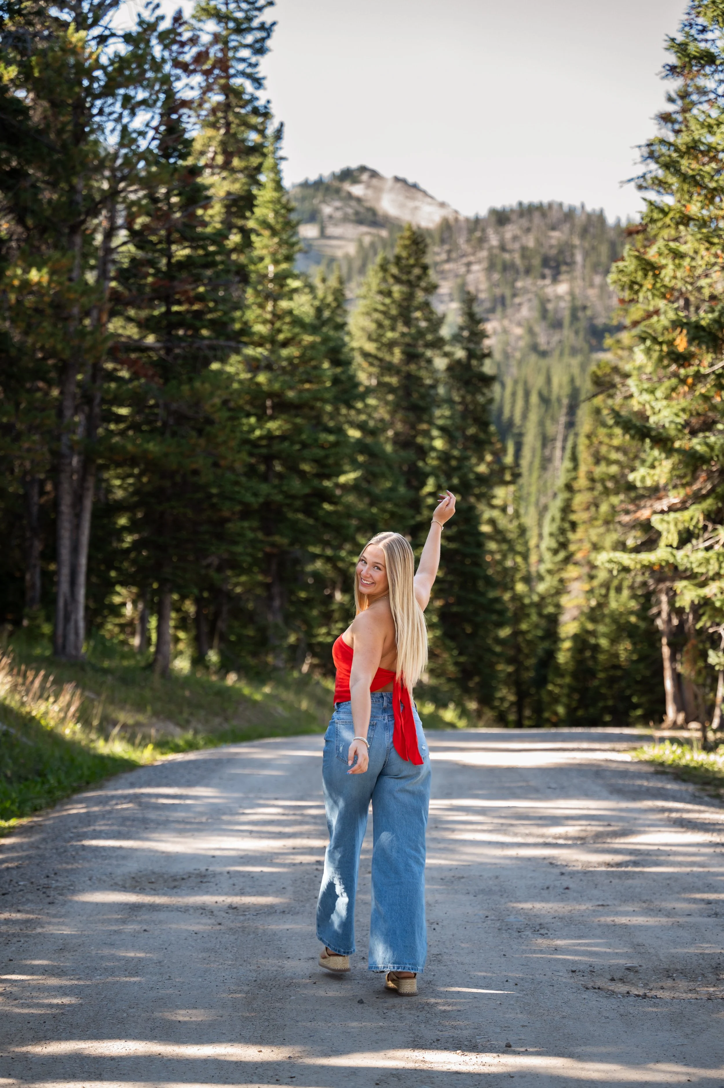 A young woman with long blonde hair smiling and waving while walking on a dirt road surrounded by tall pine trees in a forested mountainous area.