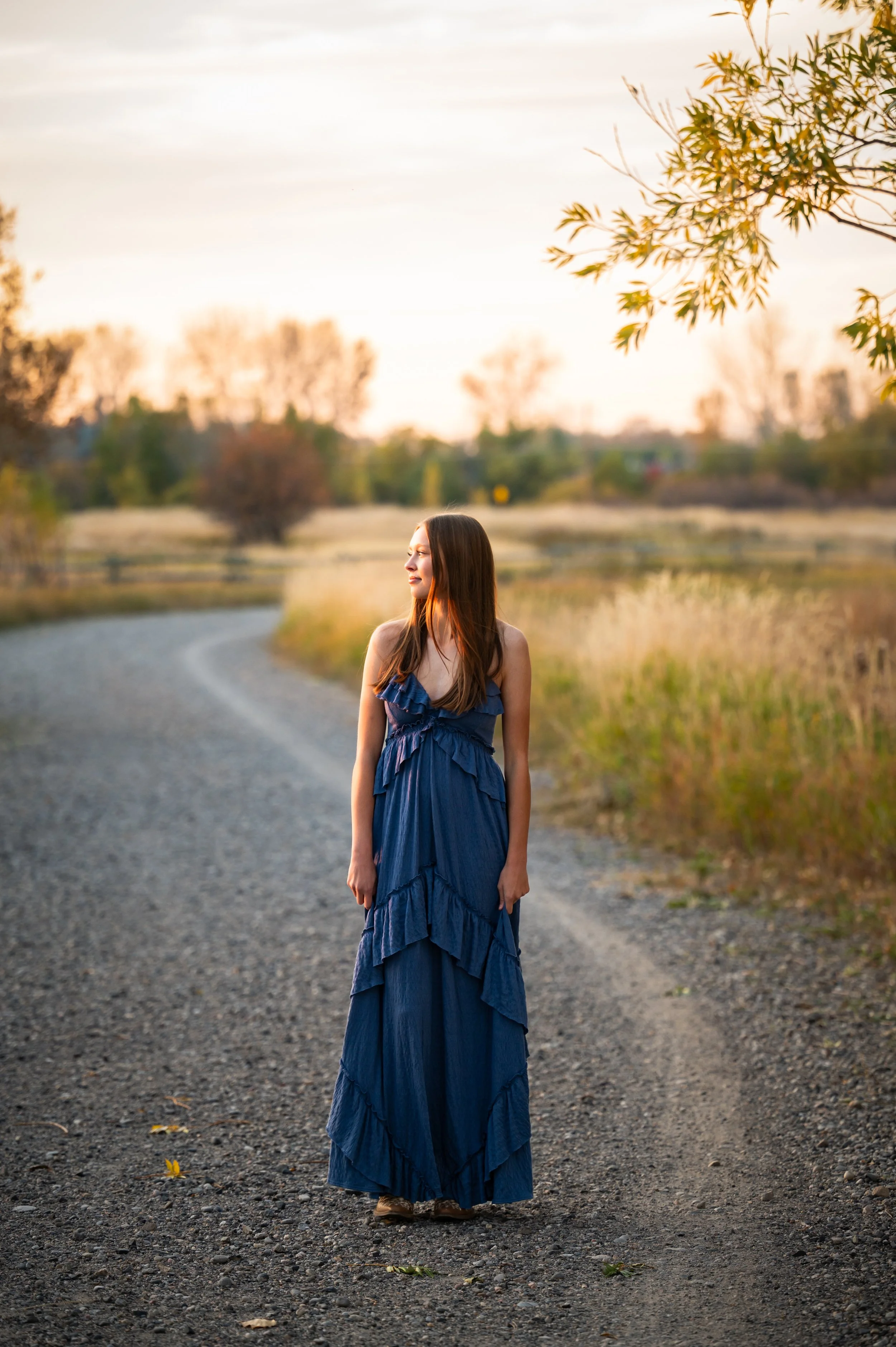 A woman in a blue dress walking on a gravel path in a rural area during sunset with trees and grassy fields in the background.