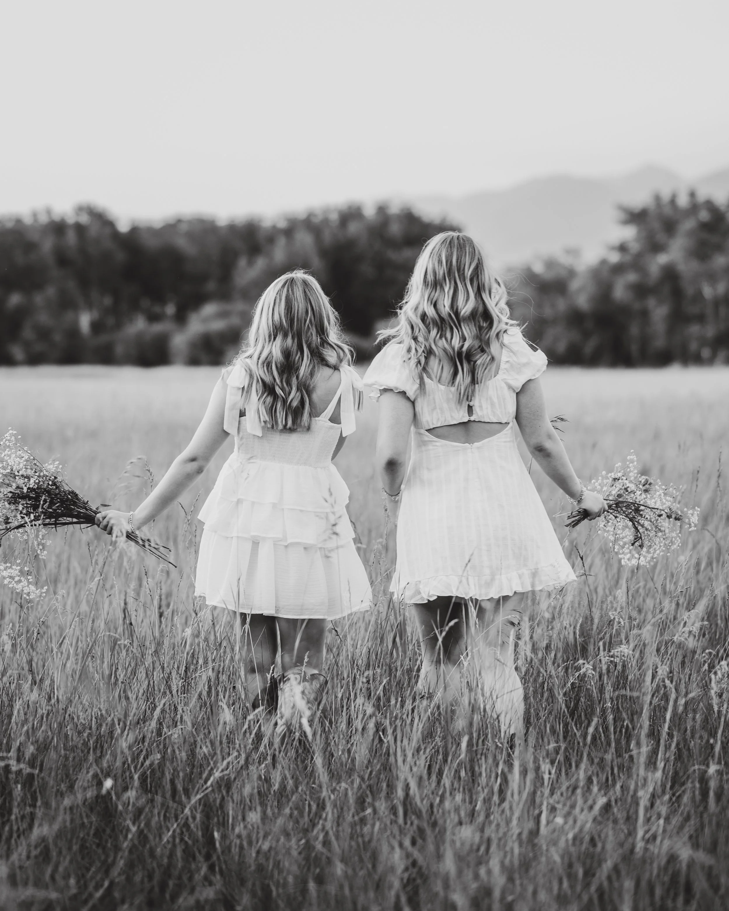 Two women walk through a grassy field, holding flowers in their hands, seen from behind, in black and white.
