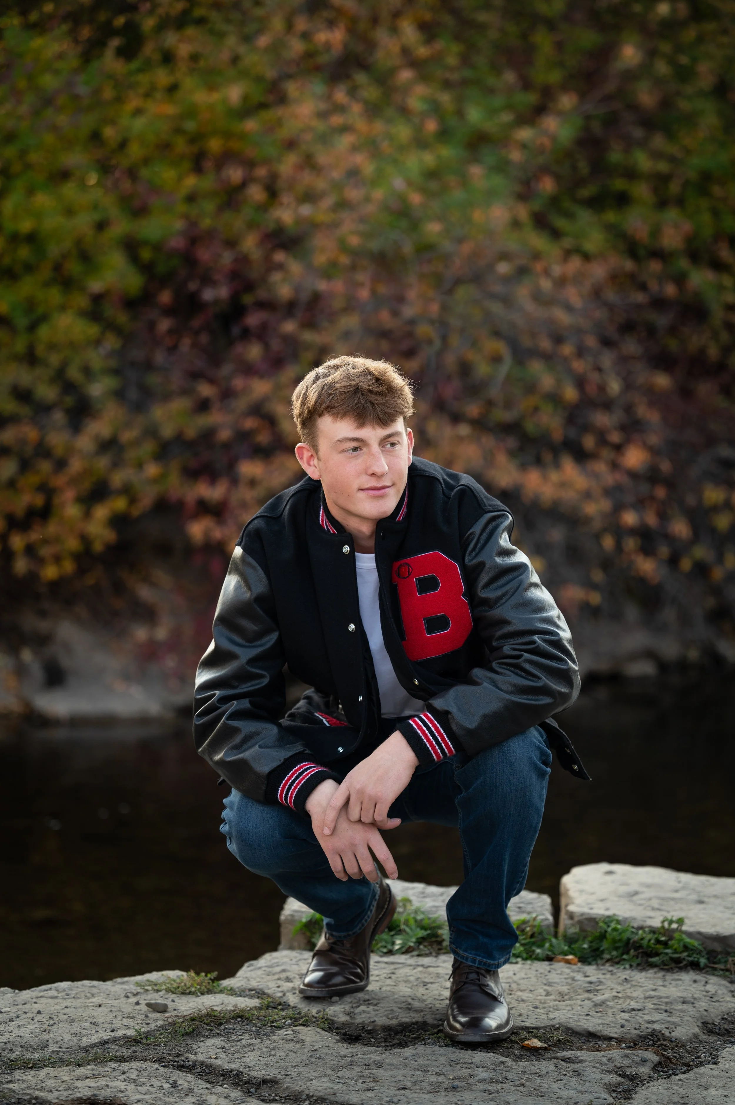 A young man in a black and gray varsity jacket with a red letter B, blue jeans, and black shoes crouches on a rock near a body of water with autumn-colored trees in the background.