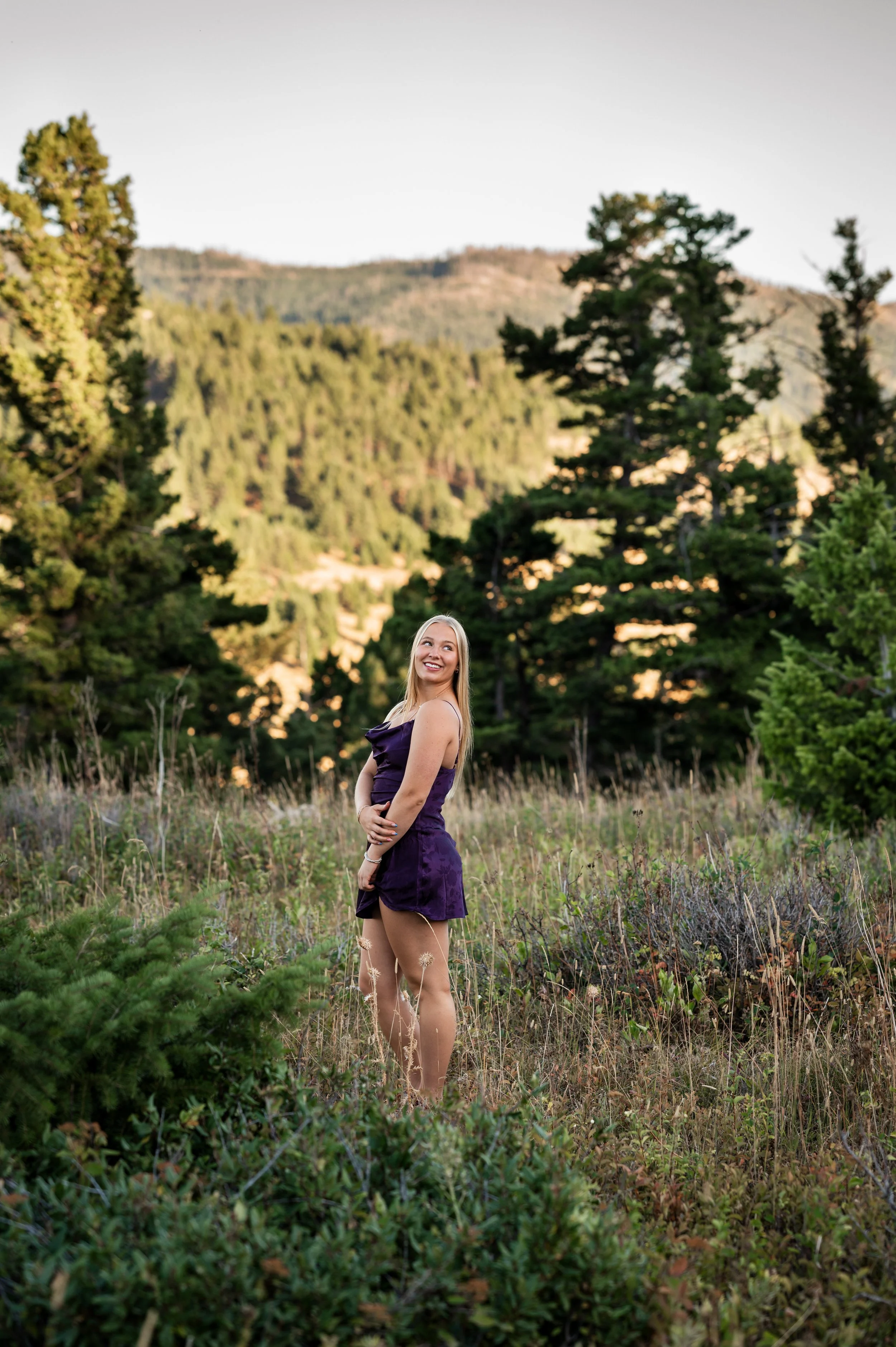 A young woman in a purple dress standing in a grassy field with trees and mountains in the background, smiling and looking upwards.