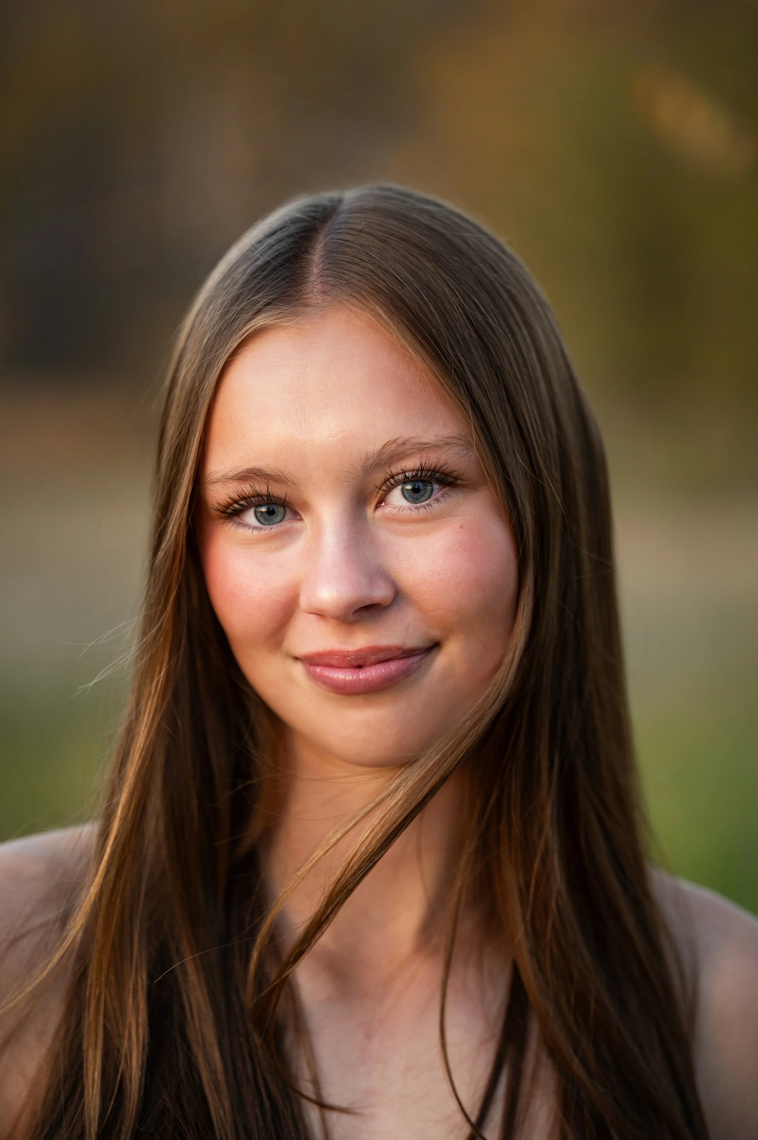 Close-up portrait of a young woman with long brown hair and blue eyes, outdoors during golden hour.