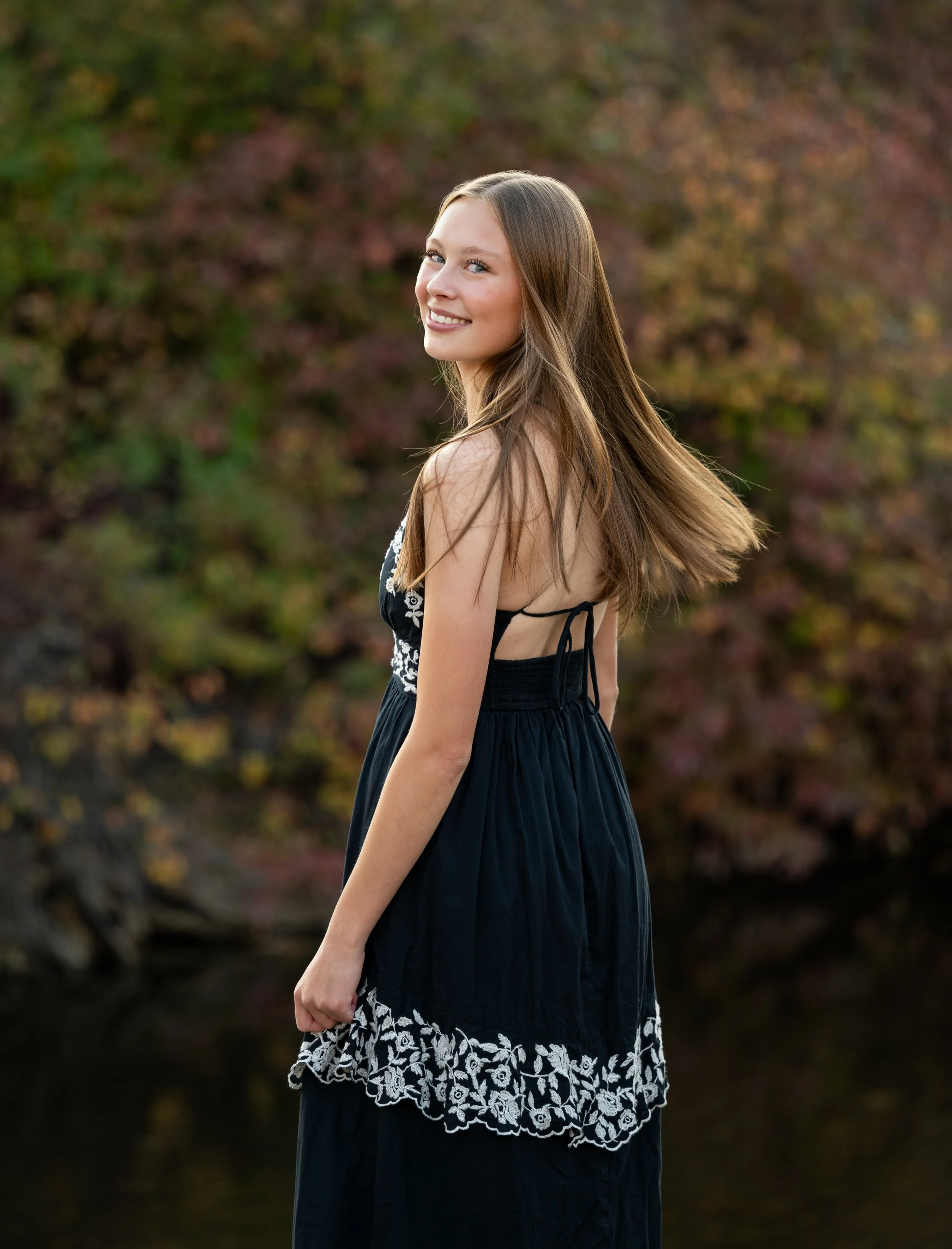 A young woman with long brown hair in a black dress with white embroidery, smiling outdoors in a park with autumn trees in the background.