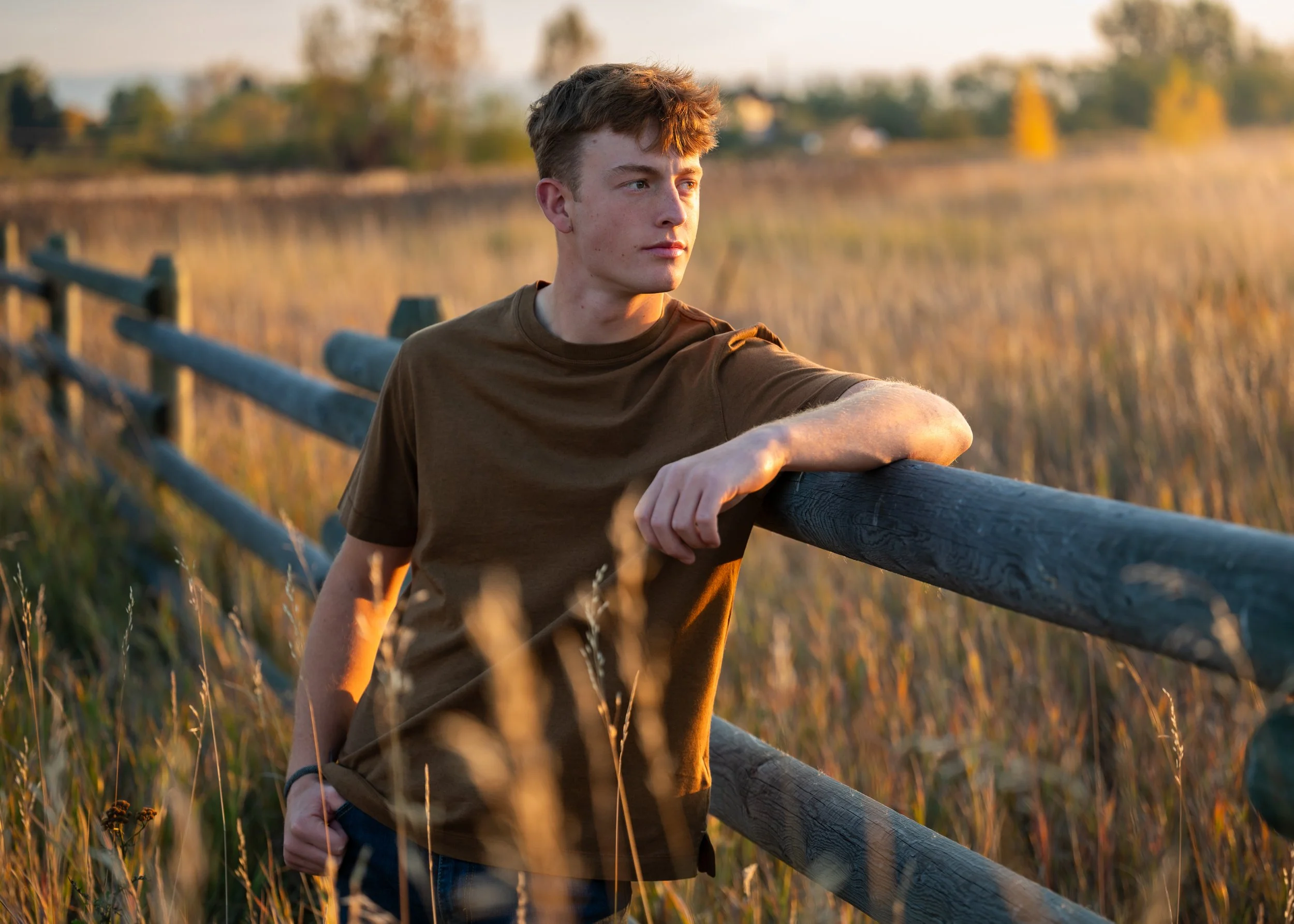 A young man in a brown t-shirt leaning on a wooden fence in a field at sunset, looking into the distance.